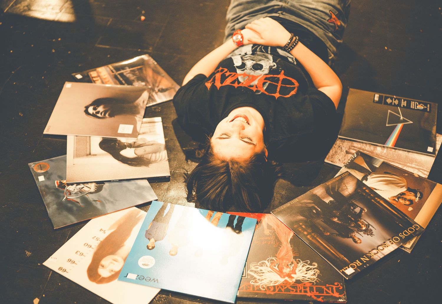 Person lying on floor surrounded by vinyl record albums, holding a camera, wearing a black hat and t-shirt.