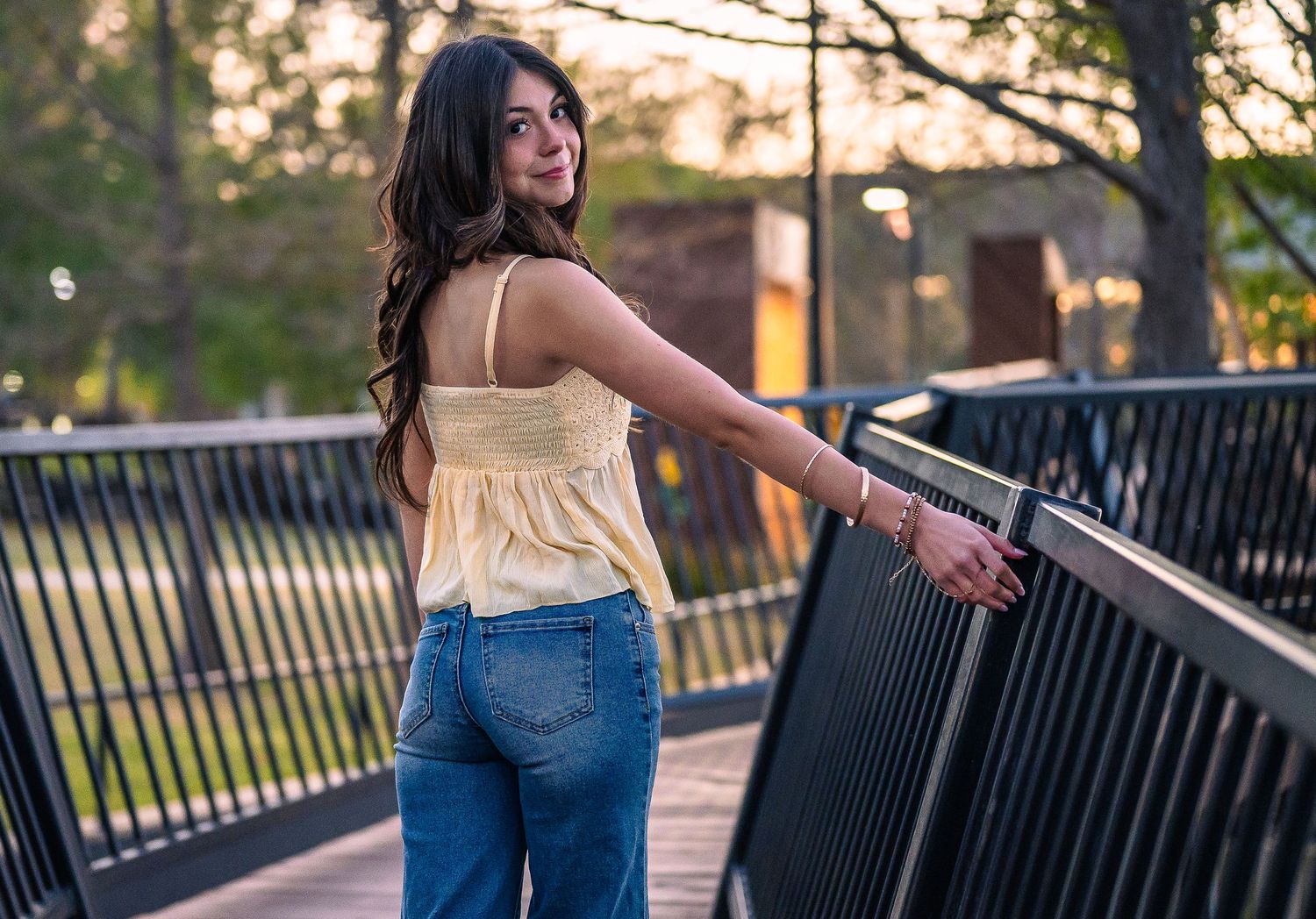 Young woman in yellow top and jeans smiling over shoulder on a bridge walkway with bokeh green trees background.