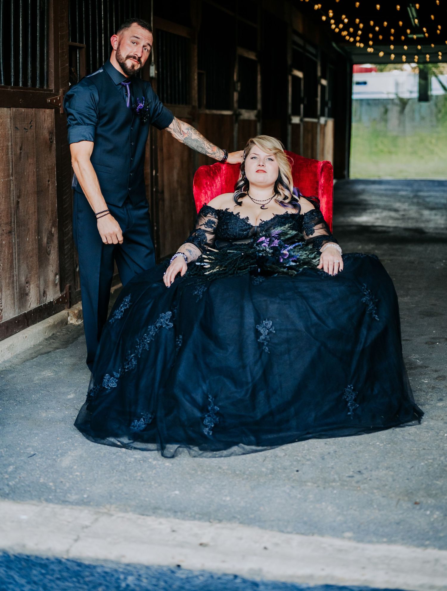 A couple sits together on a rustic wooden floor wearing formal attire before their wedding with string lights glowing in the background.