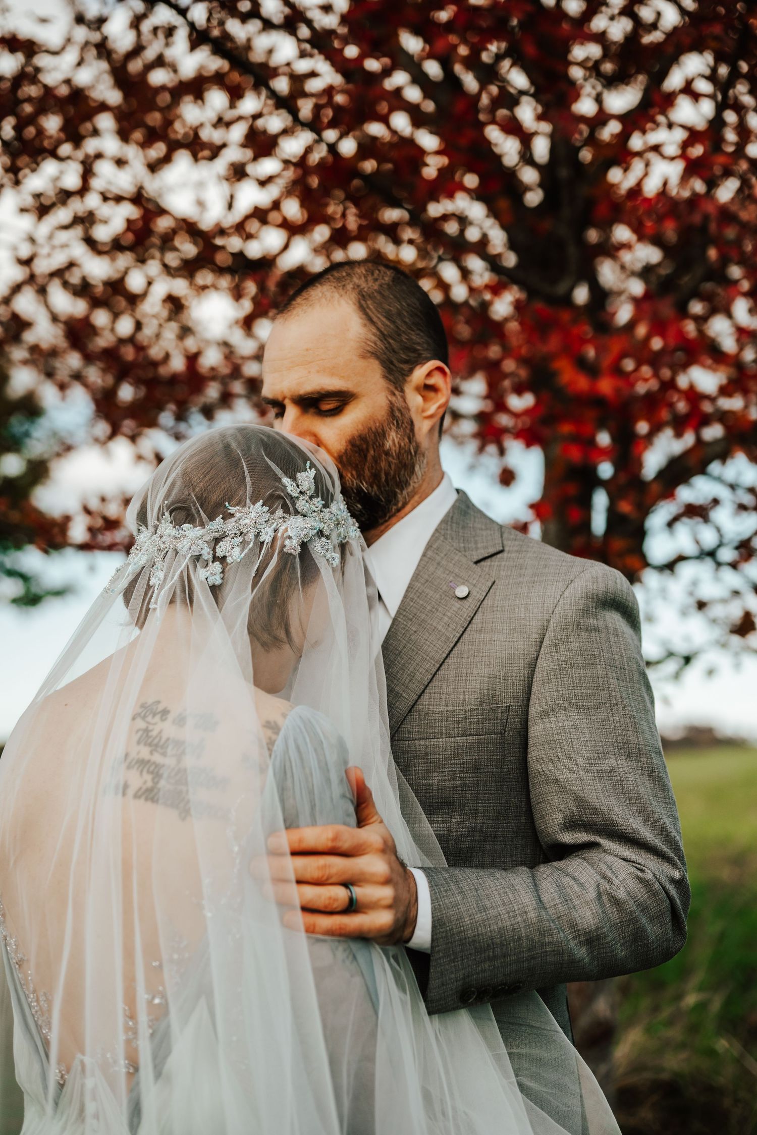 A romantic wedding moment captured under autumn trees with red foliage and a flowing white veil.