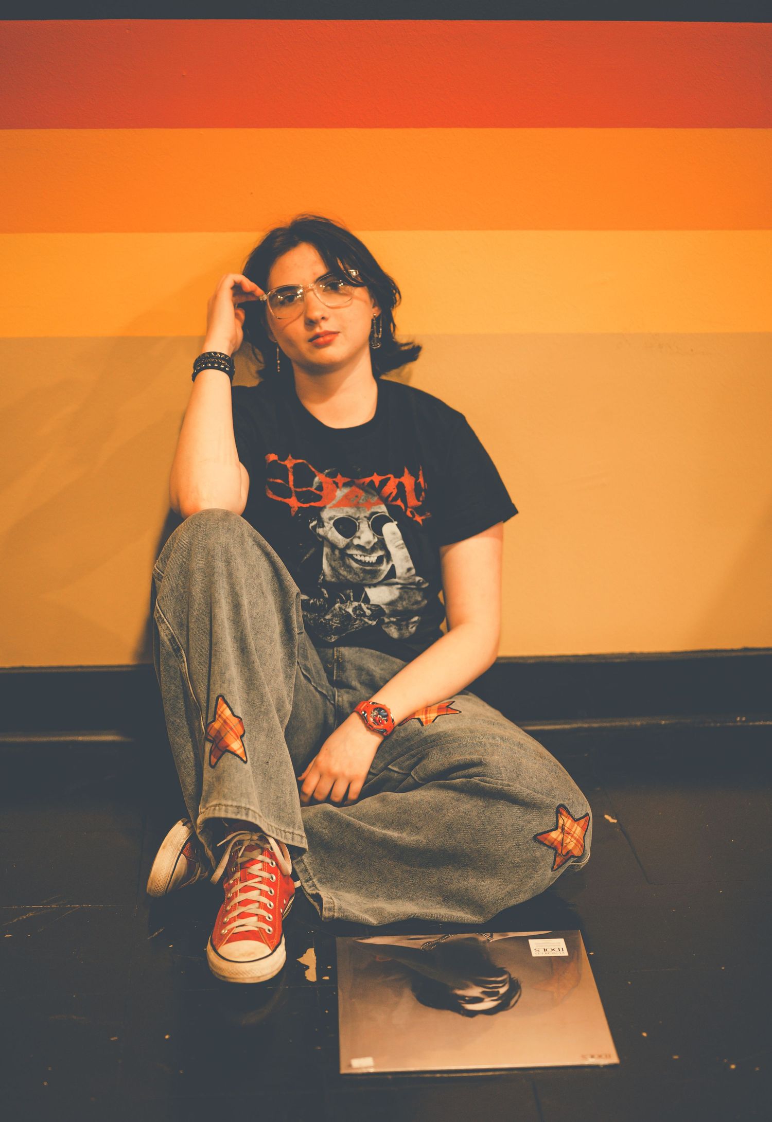 Young person in graphic tee and star-patched jeans sits against colorful orange striped wall wearing red Converse sneakers.