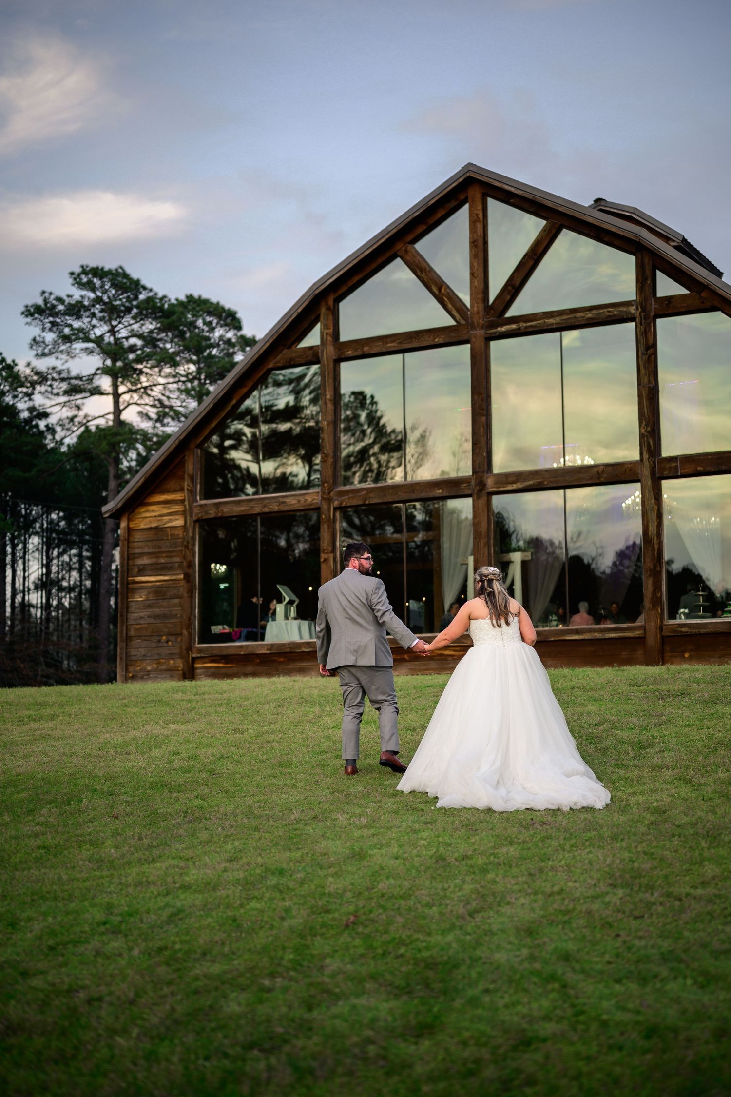 Bride and groom holding hands walking toward a rustic wooden barn wedding venue with large glass windows at dusk.