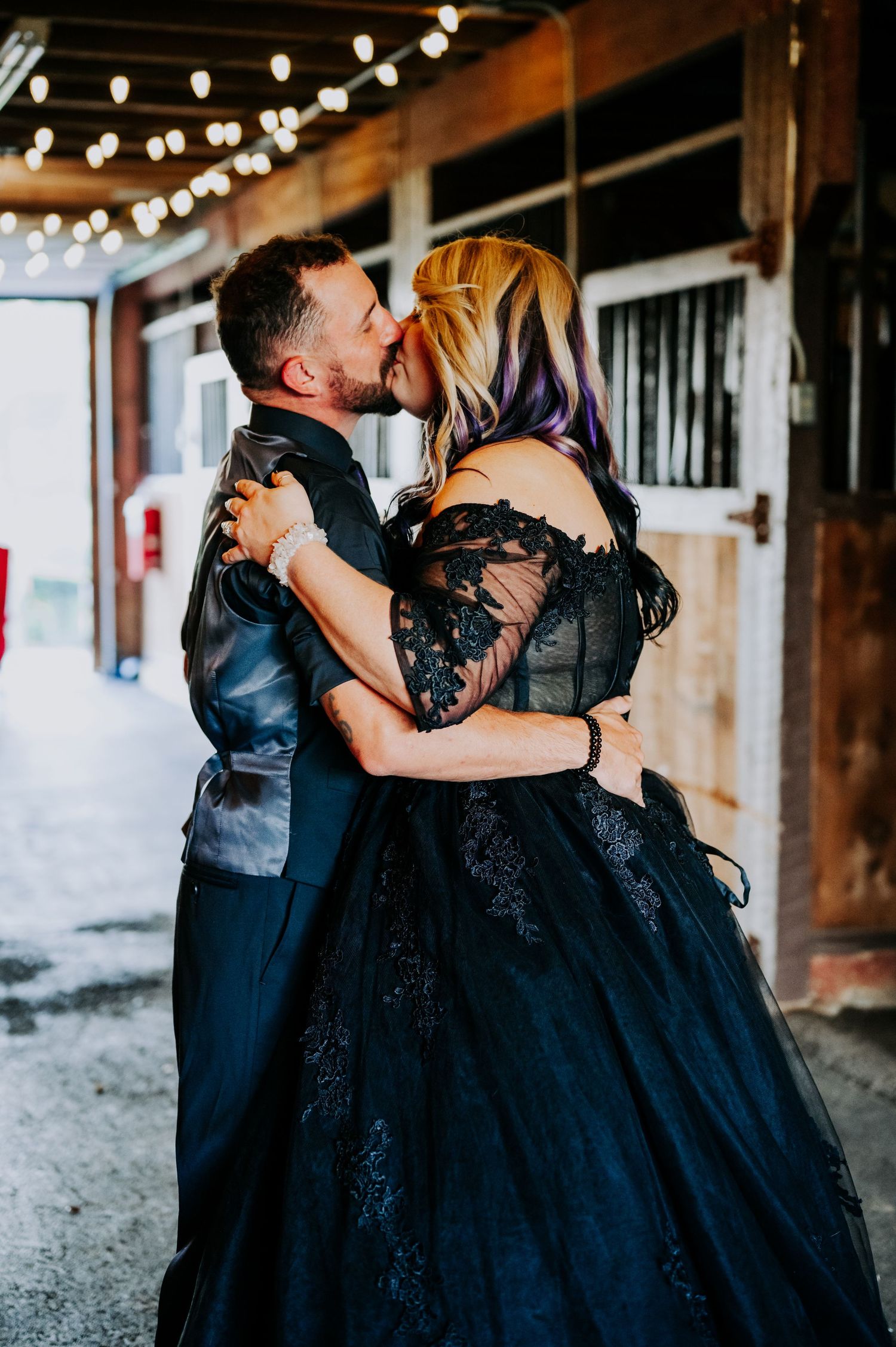 Couple in black formal wear embracing under string lights in a rustic venue.