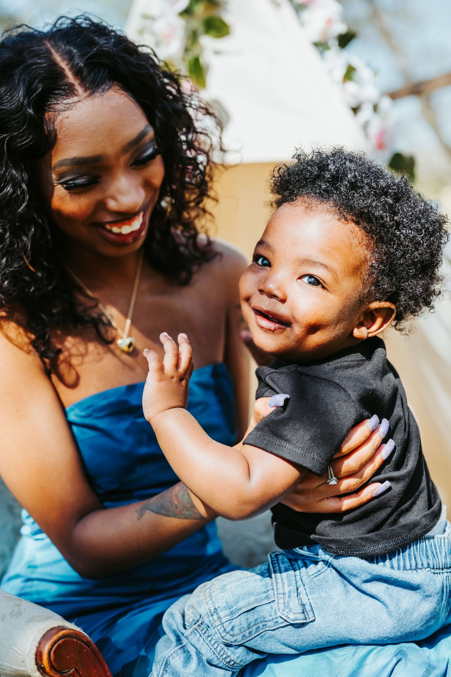 A smiling Black mother in a blue dress holds her happy toddler son outdoors in a bright, floral setting.