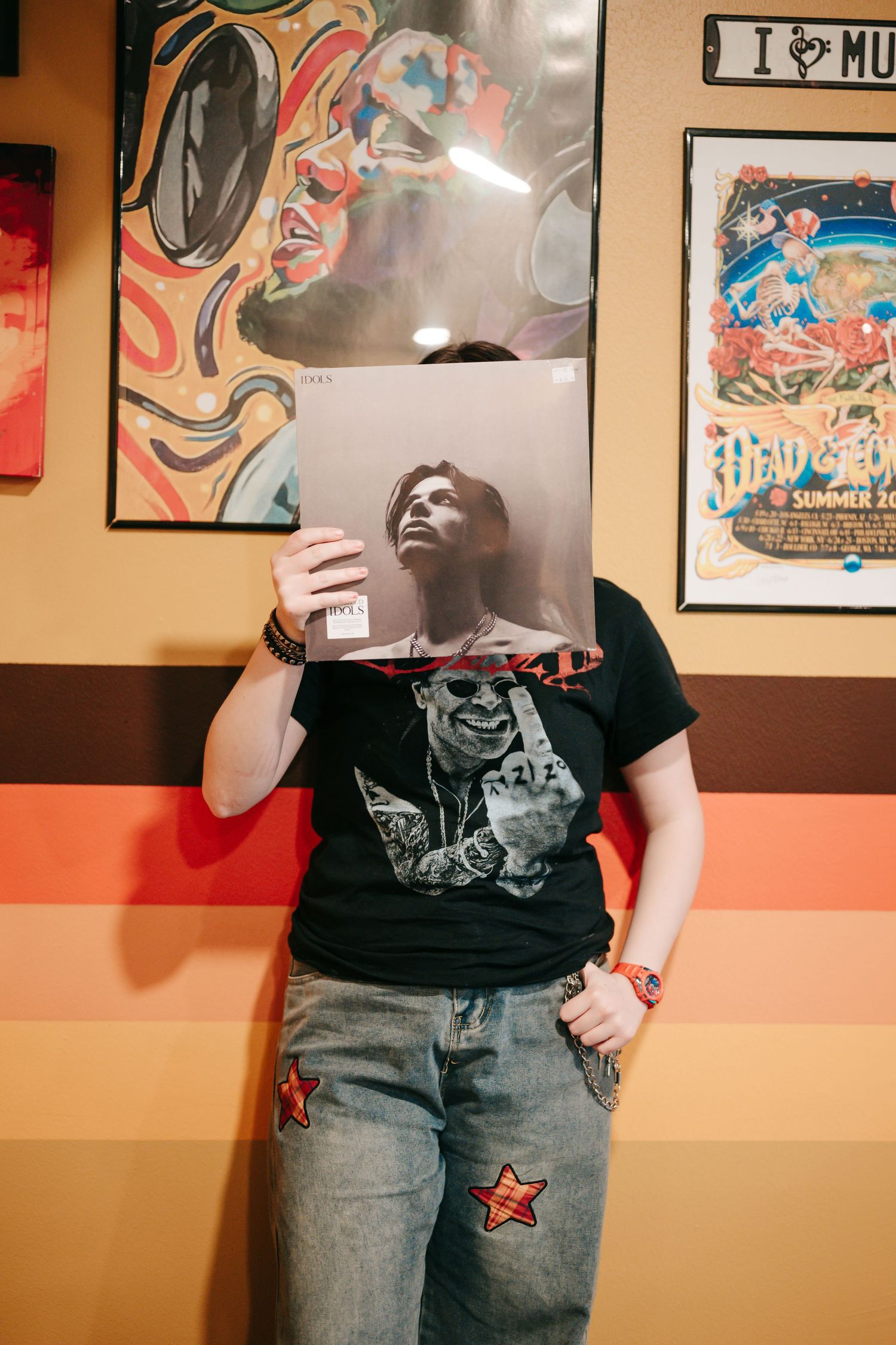 Person holding vinyl record cover in front of face, wearing graphic tee and star-patched jeans in a music store.