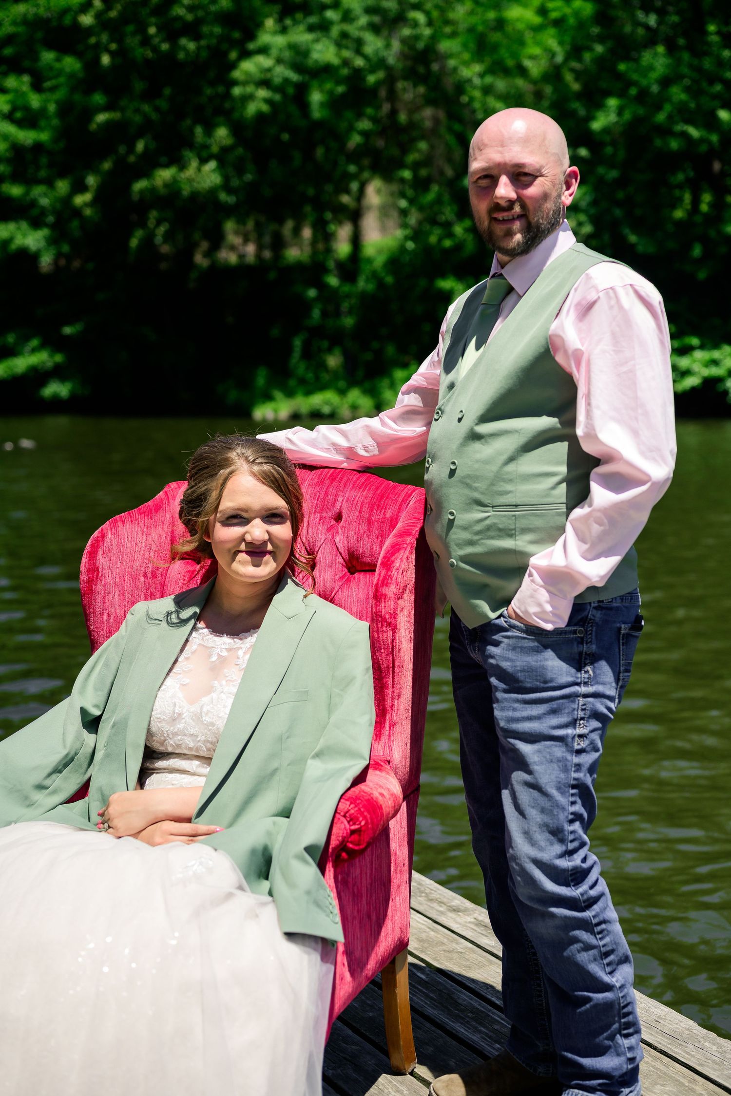 Bride in green jacket sits in red velvet chair beside groom in vest on wooden dock near serene lake.