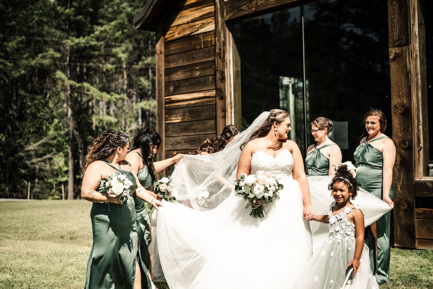 Bride in white gown and veil surrounded by bridesmaids in sage green dresses outside rustic wooden chapel.