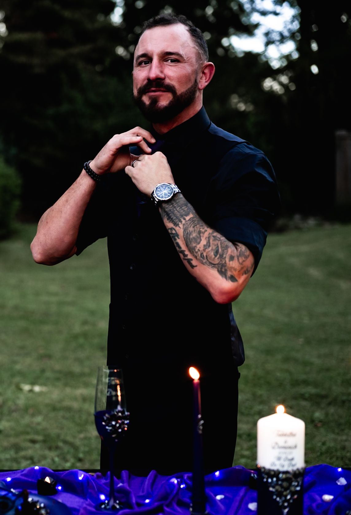 Man in black shirt with tattoos posing near candlelit purple table settings outdoors.