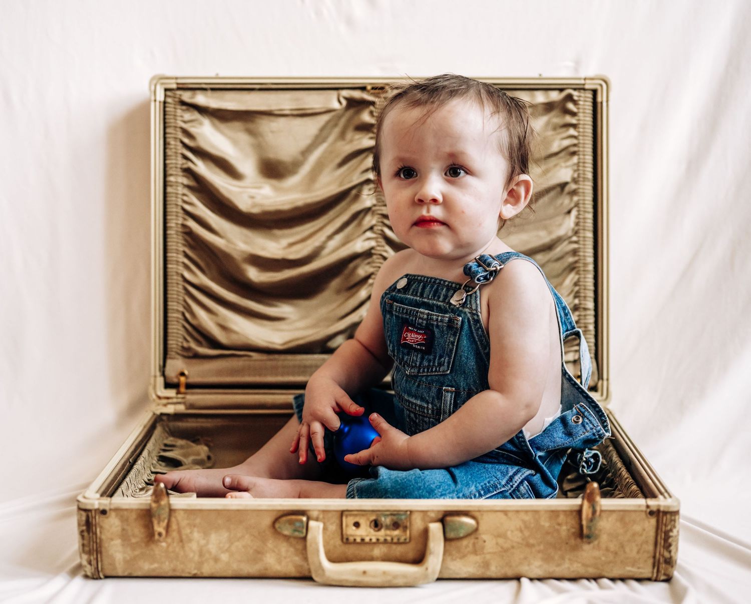 A baby in denim overalls peeks out from inside a vintage suitcase in a creative photo series.
