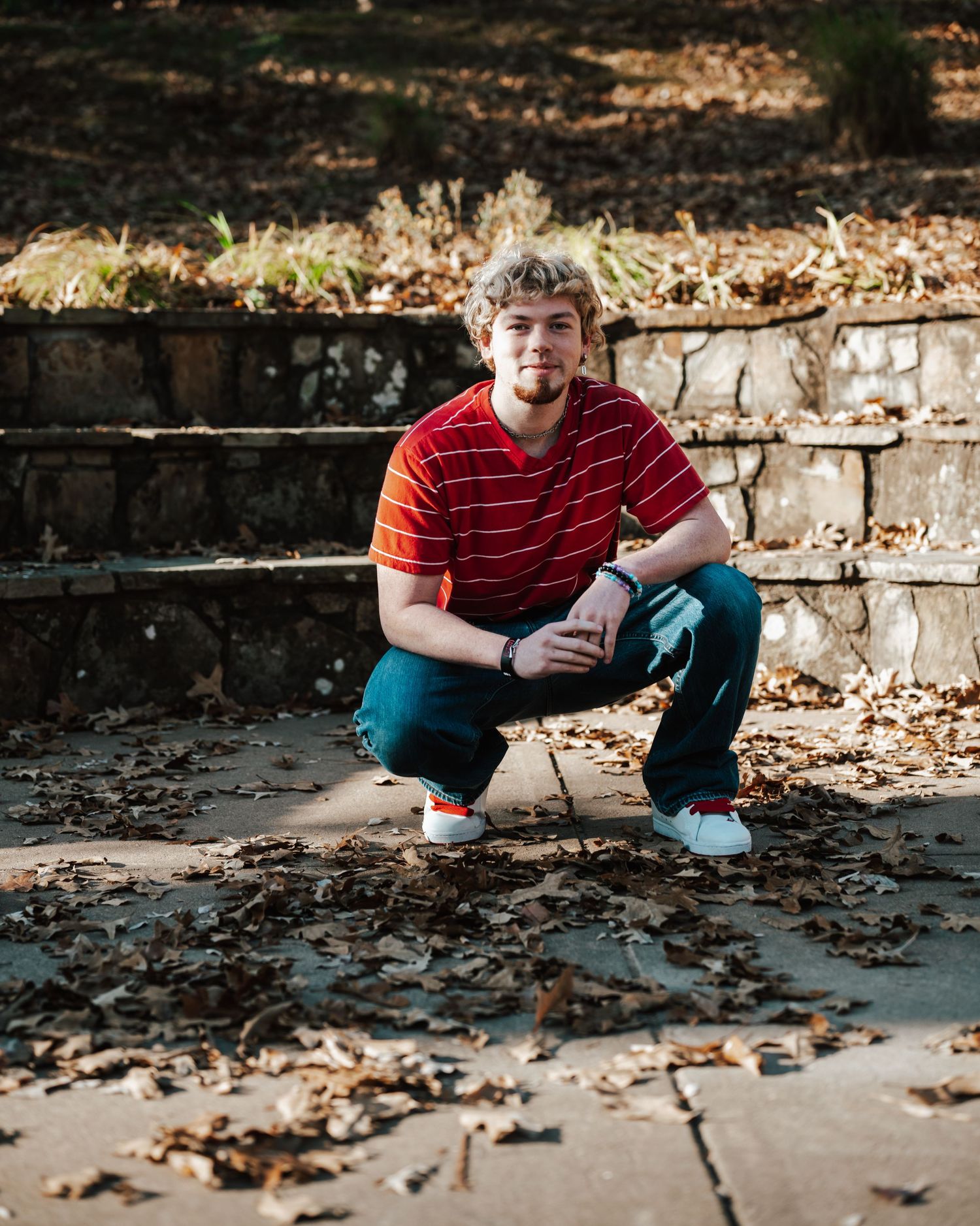 Young man in red striped shirt crouching on stone steps surrounded by fallen autumn leaves outdoors.