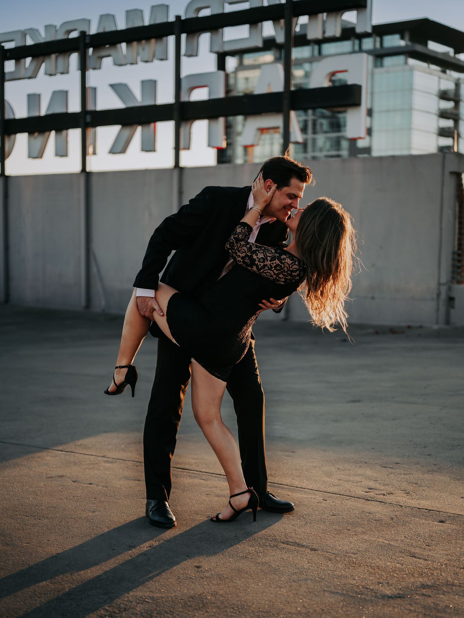 A couple in black outfits share a romantic dip kiss on a rooftop at golden hour.