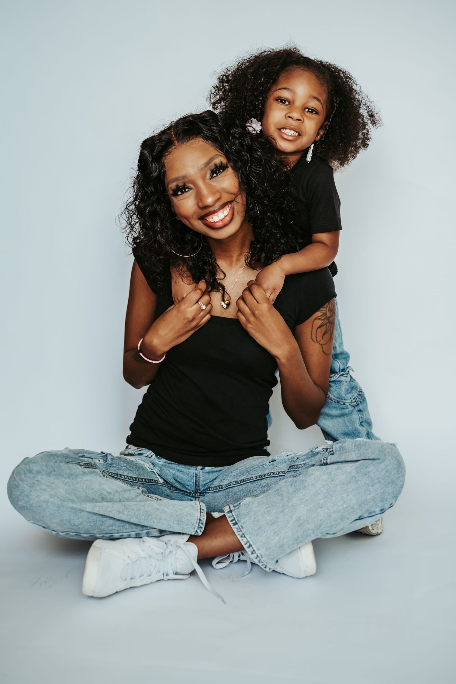 A smiling woman and young girl in matching black tops and jeans pose together on a white background.