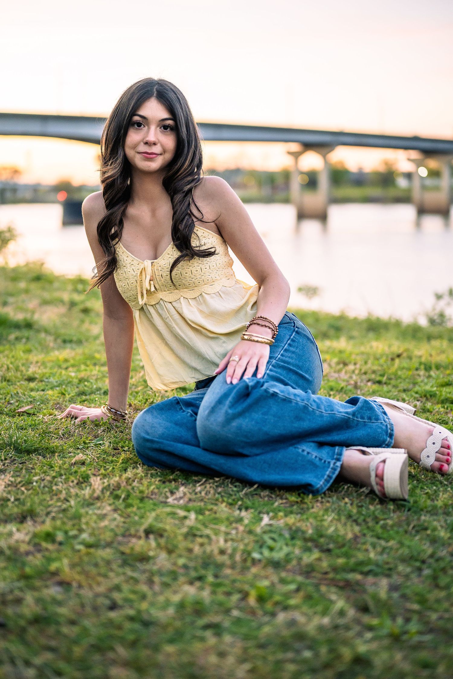 Young woman in yellow top and jeans sitting on grass by a river at sunset.
