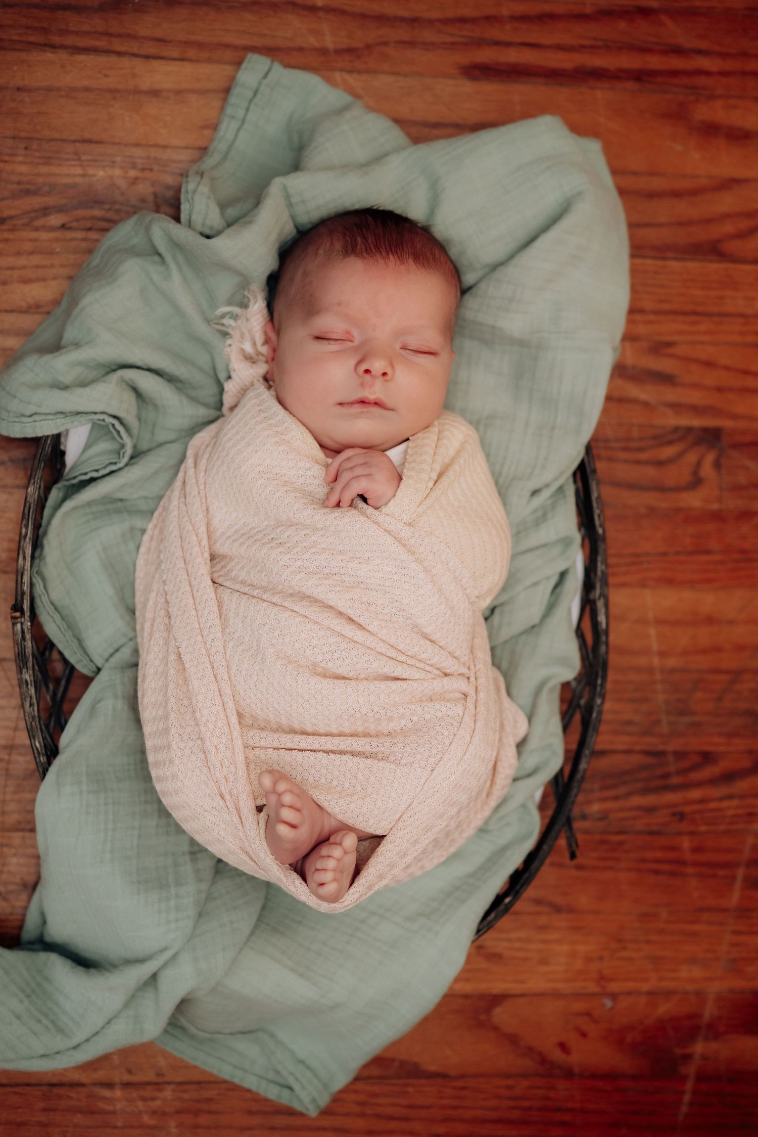 Sleeping newborn wrapped in soft pink swaddle blanket lying on sage green fabric against wooden floor.
