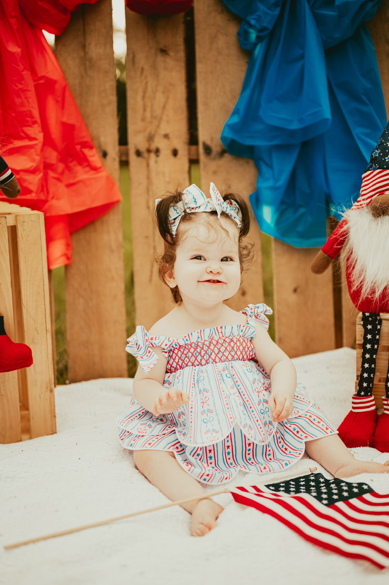 Baby in patriotic outfit posing with American flag against wooden backdrop.