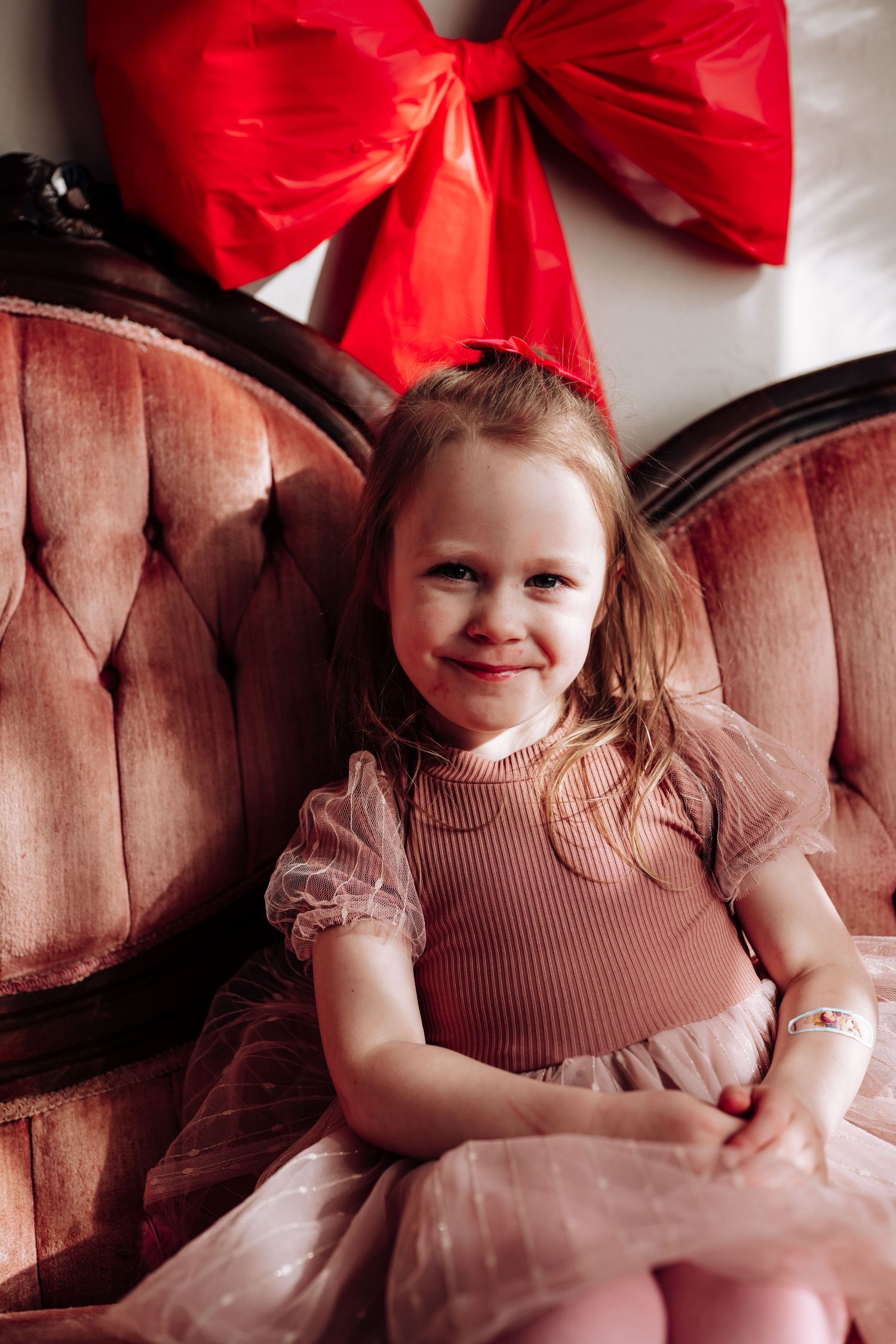 Young girl in pink tutu dress smiling on vintage velvet chair with red bow decoration.