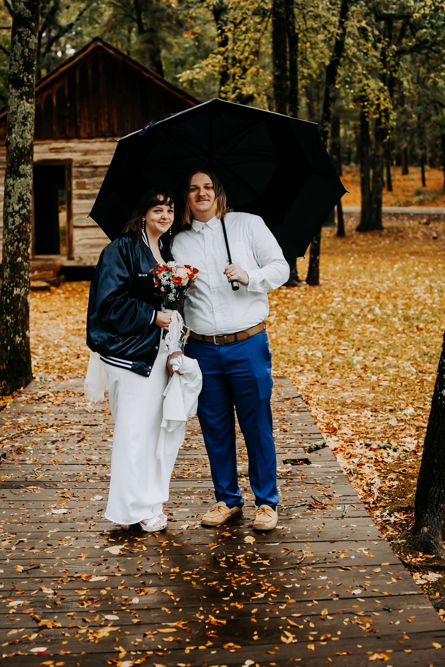 A couple shares an intimate moment under an umbrella on a path covered in fallen autumn leaves.