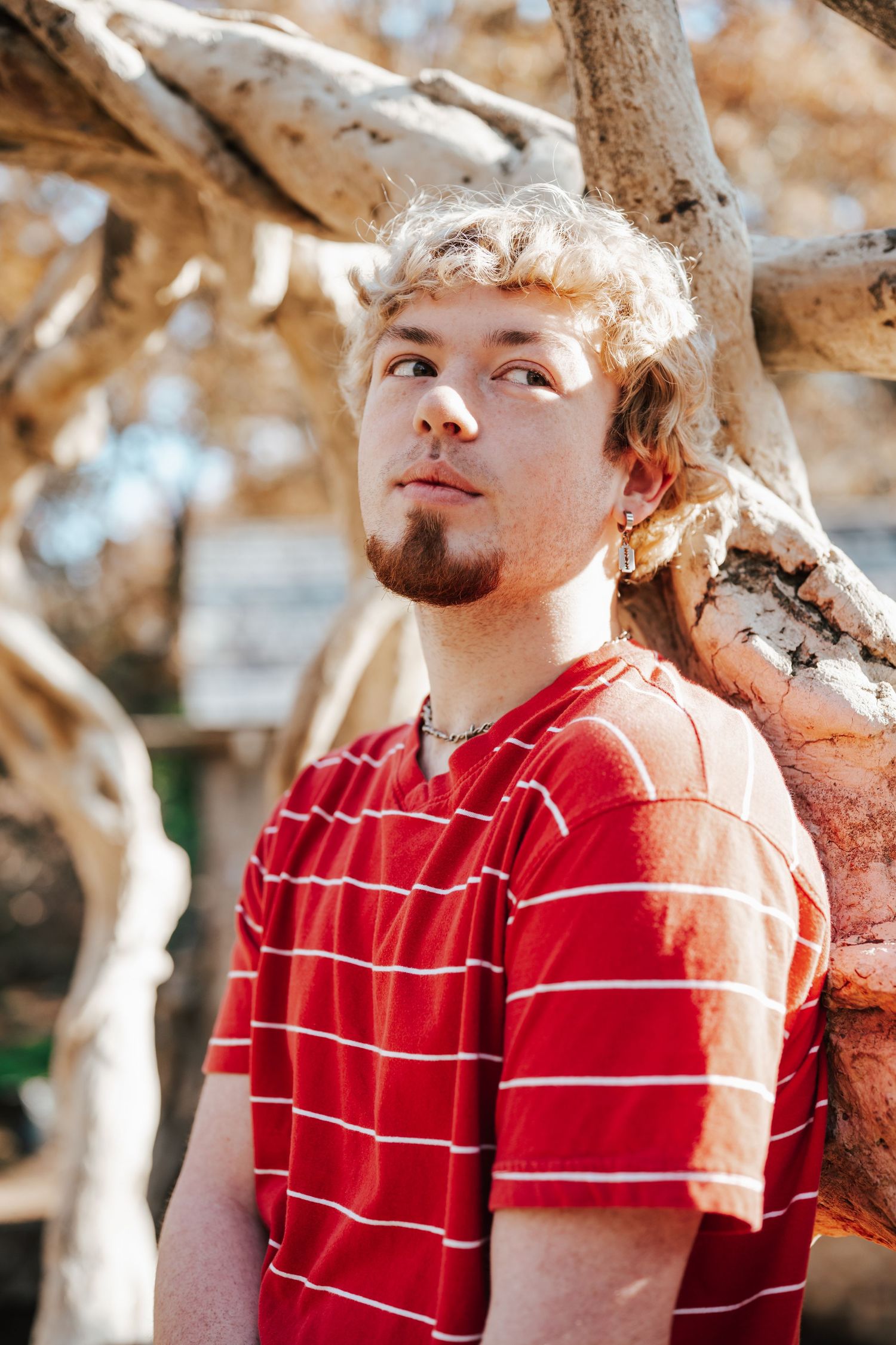 Young man with blond curly hair and beard wearing red striped t-shirt leaning against tree branches outdoors.