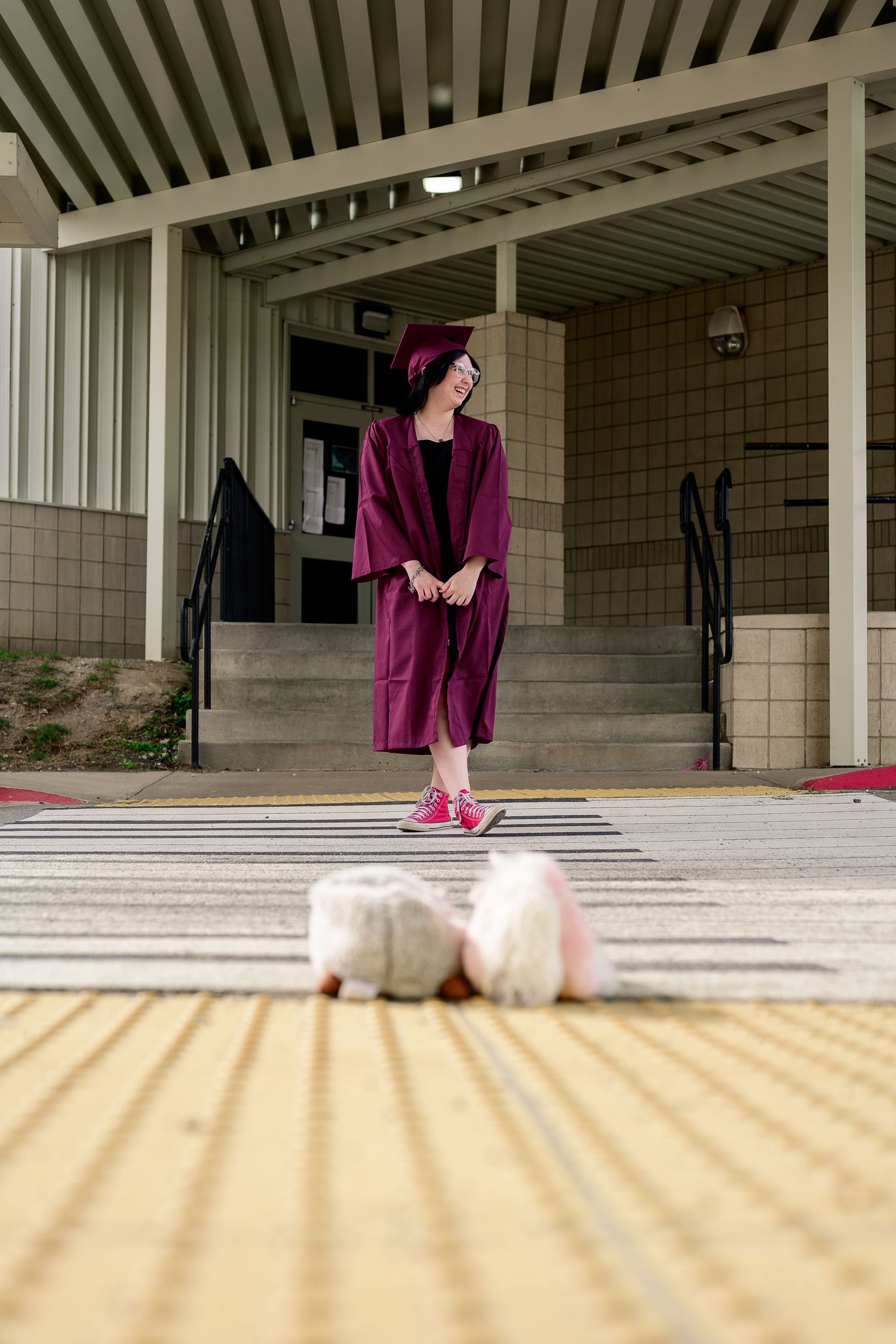 Graduate in burgundy cap and gown with pink sneakers poses outside school building during graduation photo session.