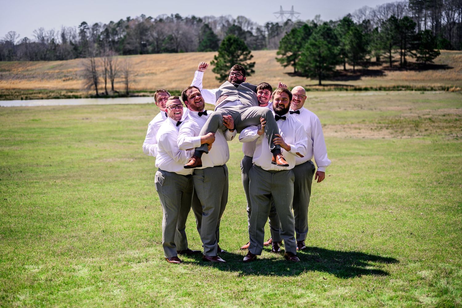 Groomsmen in gray suits lifting groom outdoors on sunny day at rustic countryside wedding venue.
