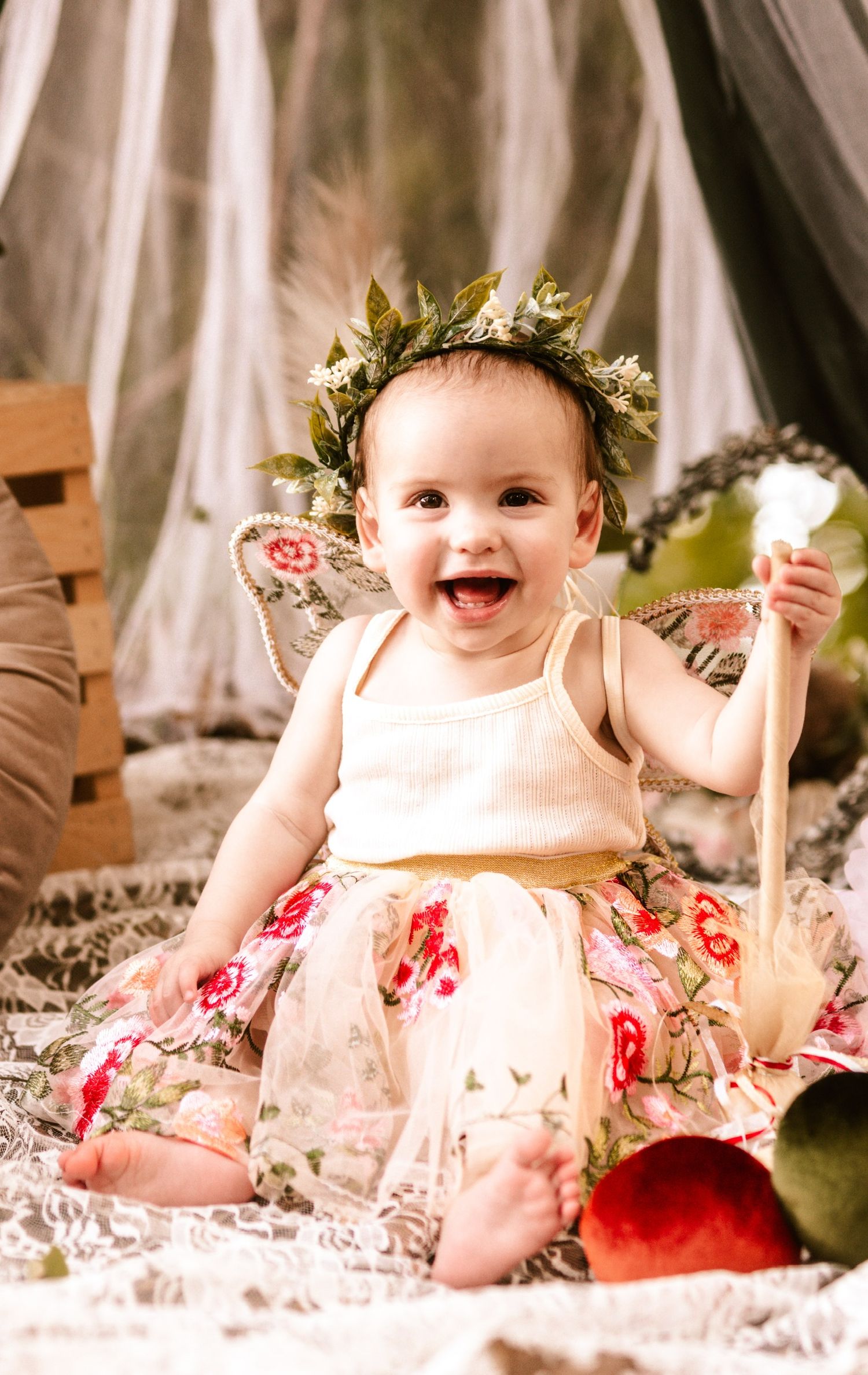 Laughing baby with leaf crown and floral dress on cream colored blanket.
