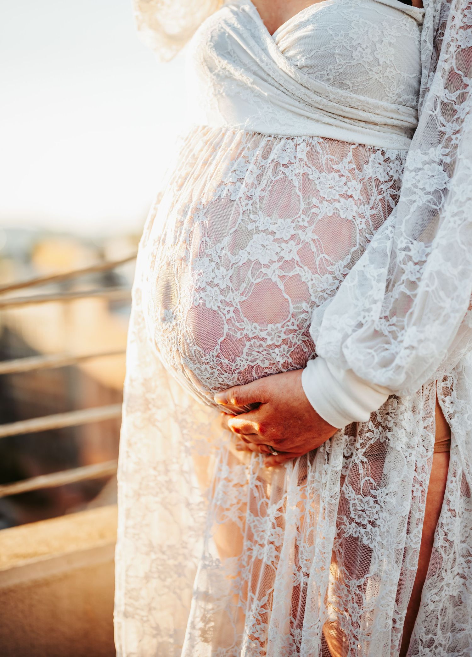 Pregnant woman in elegant white lace maternity gown cradling her baby bump outdoors in golden sunlight.