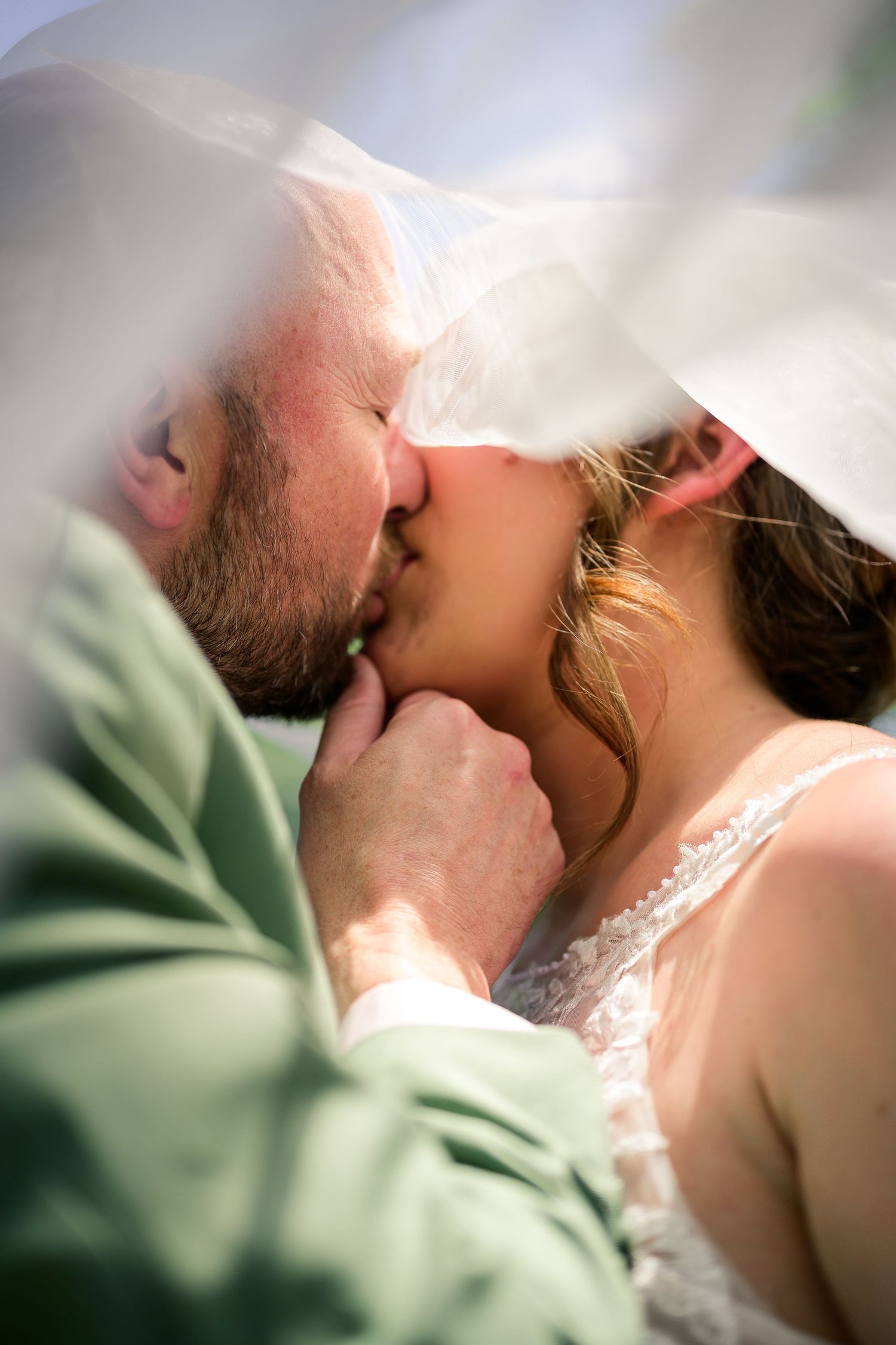 A bride and groom share a romantic kiss under a flowing white veil on their wedding day.
