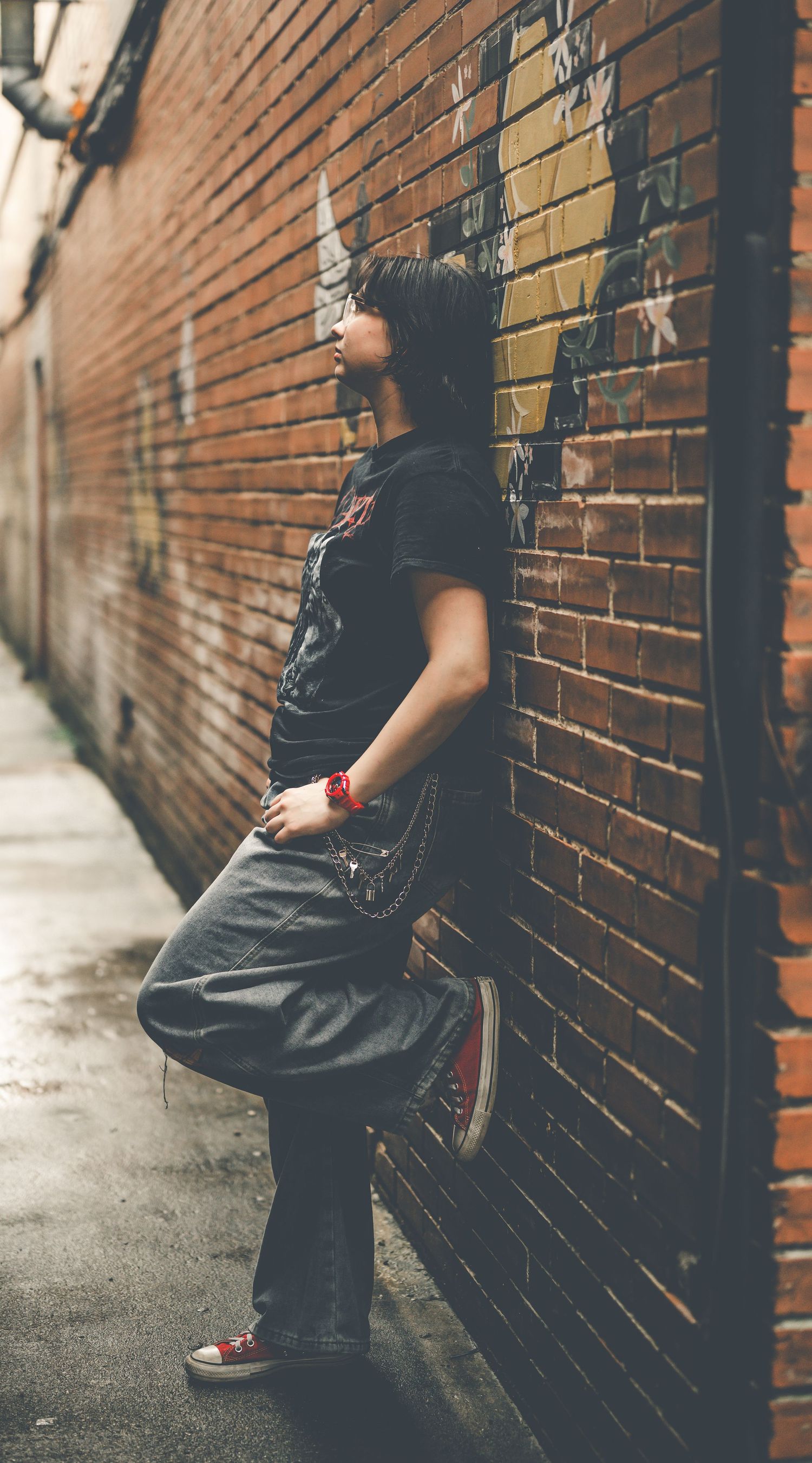 Young person leaning against a graffiti brick wall in an urban alley, wearing a black tee, gray pants, and red Converse.