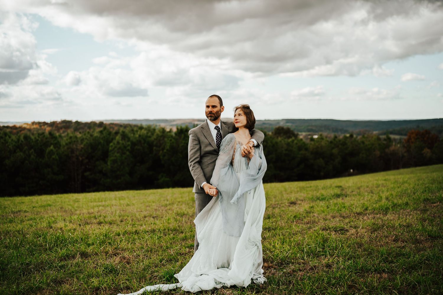 A bride and groom embraces in a scenic meadow with rolling hills and dramatic cloudy skies in the background.