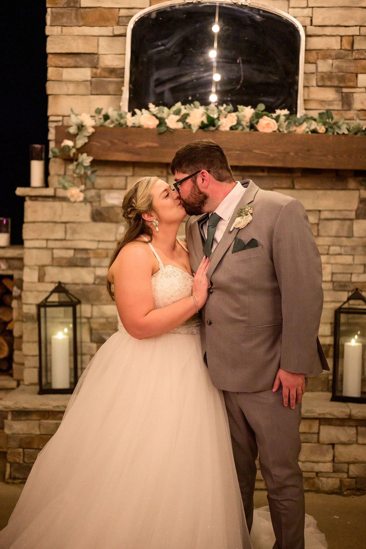 Bride and groom share a kiss in front of a stone fireplace adorned with candles and floral garland at their wedding reception.