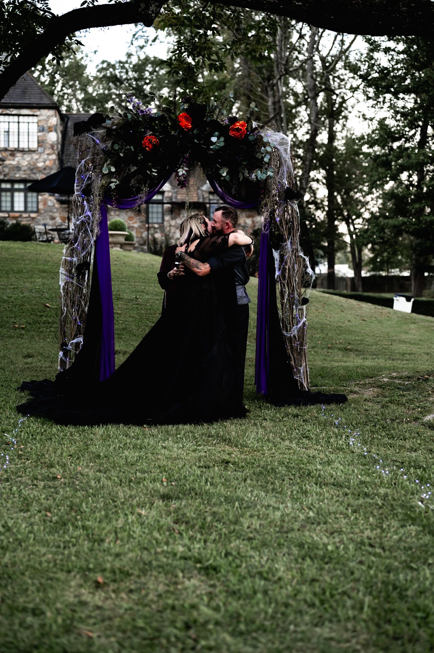 A couple in black gothic wedding attire embraces under a decorated arch with red lights on a grassy hillside.