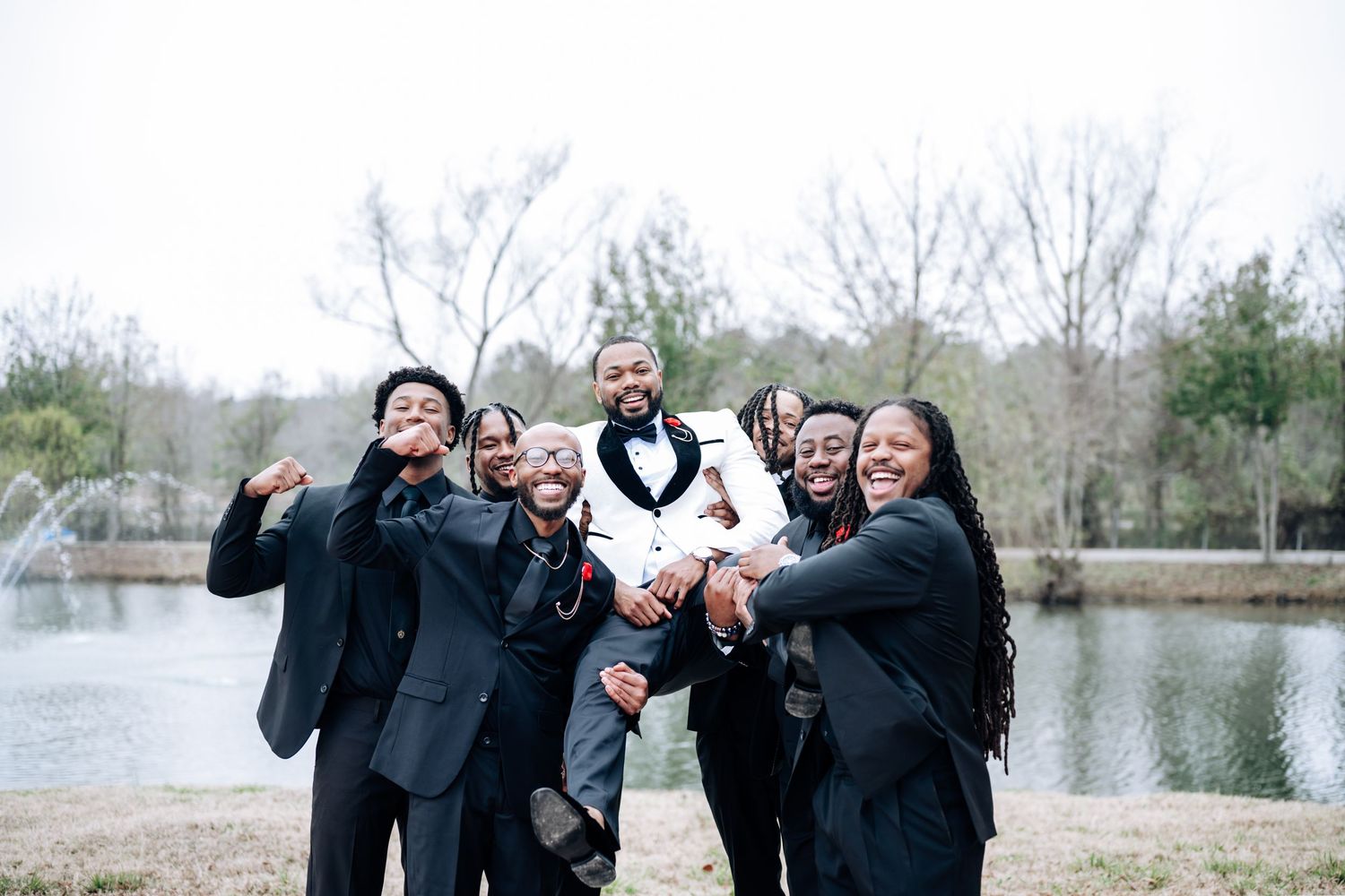 A group in formal attire poses together outdoors near a lake during a photo session.
