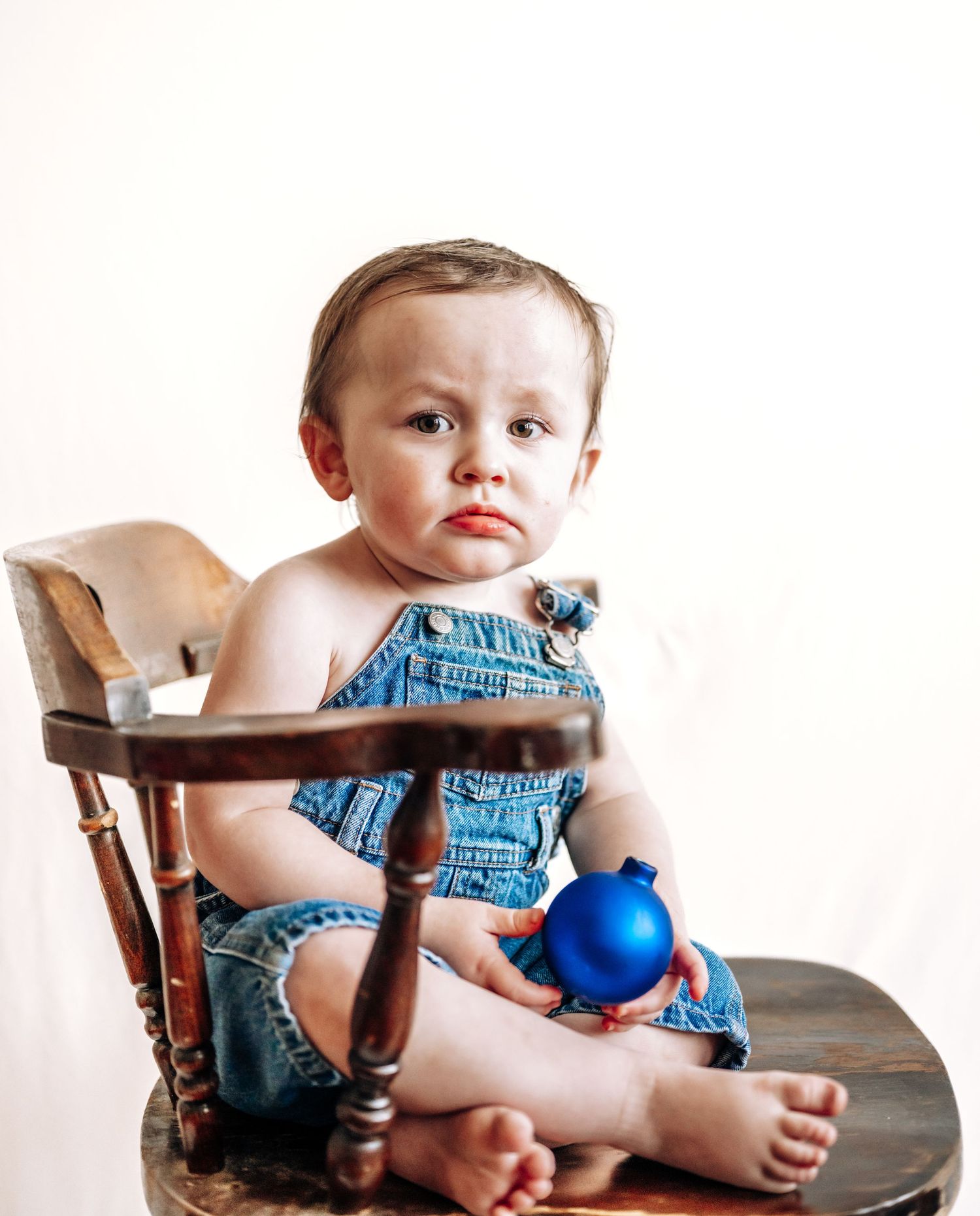 Small child wearing denim overalls sits in wooden chair holding a bright blue Christmas ornament.