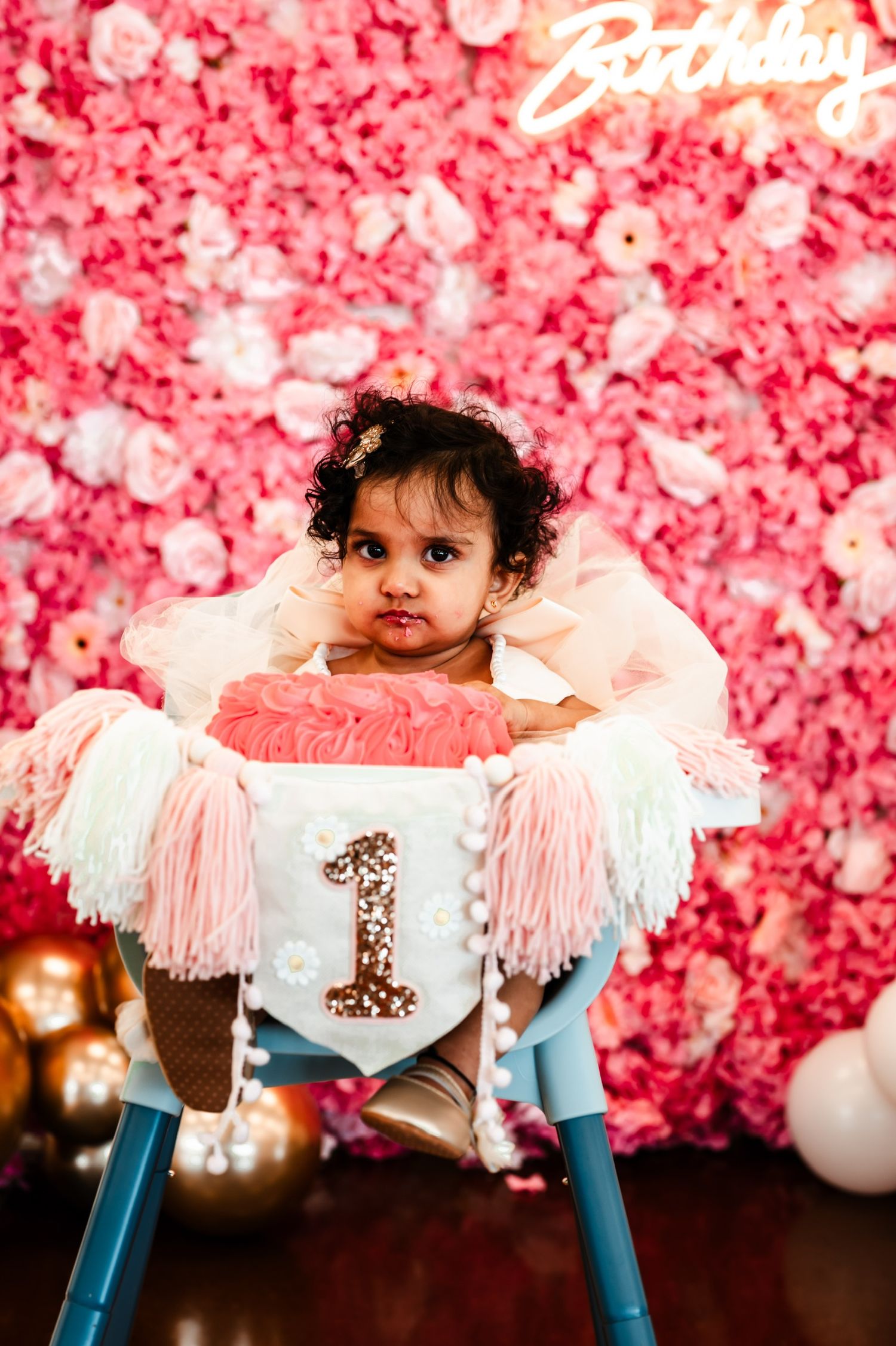 Baby sits in decorated high chair against pink floral backdrop at first birthday celebration.