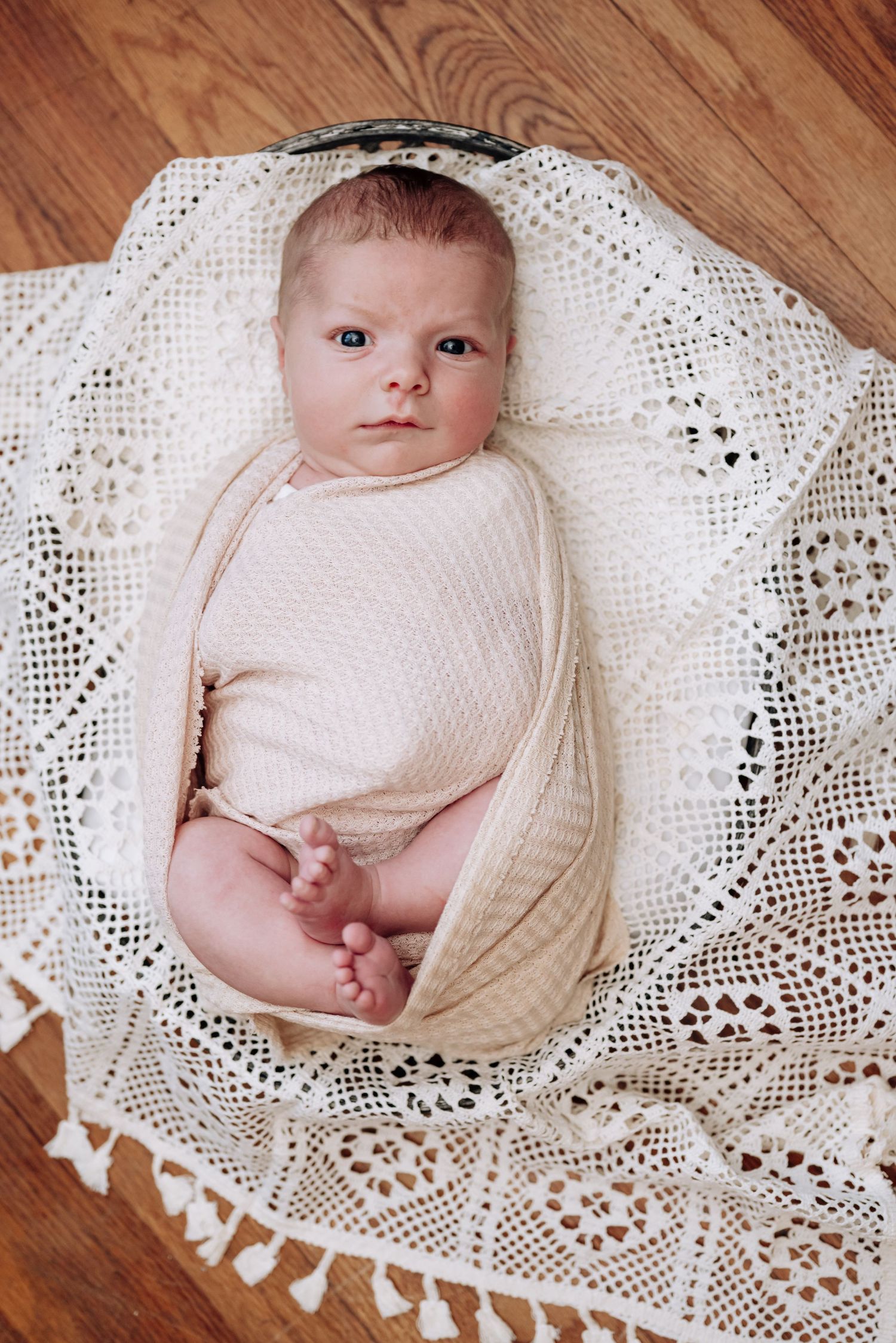 Newborn baby swaddled in cream wrap lying on white crochet blanket, looking up with alert blue eyes.