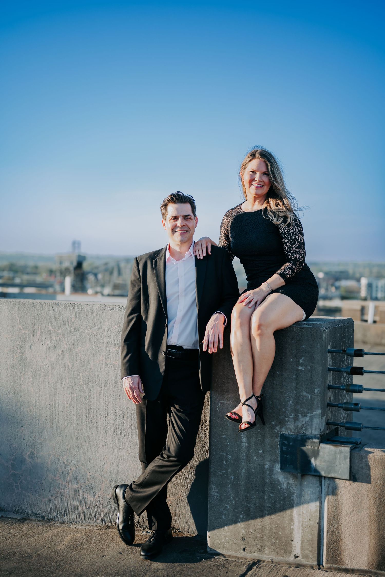 Couple posing on rooftop, man in black suit and woman in black dress, smiling against blue sky backdrop.