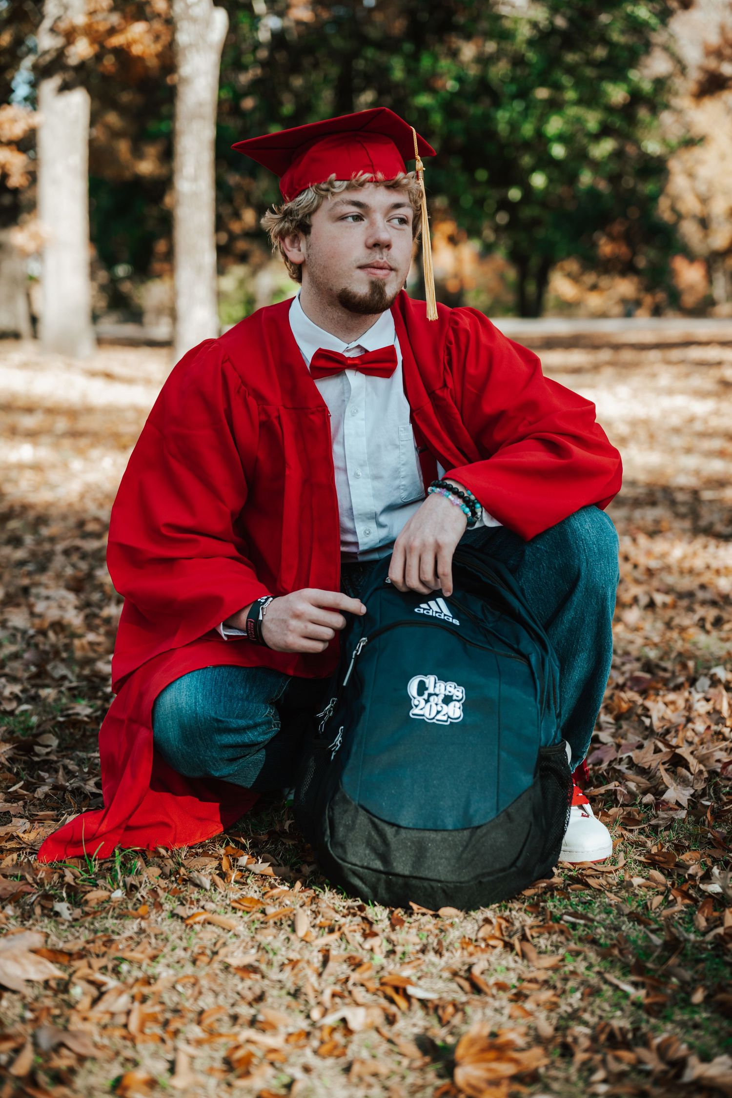 Young man in red graduation cap and gown sits outdoors on backpack among autumn leaves, wearing bow tie.