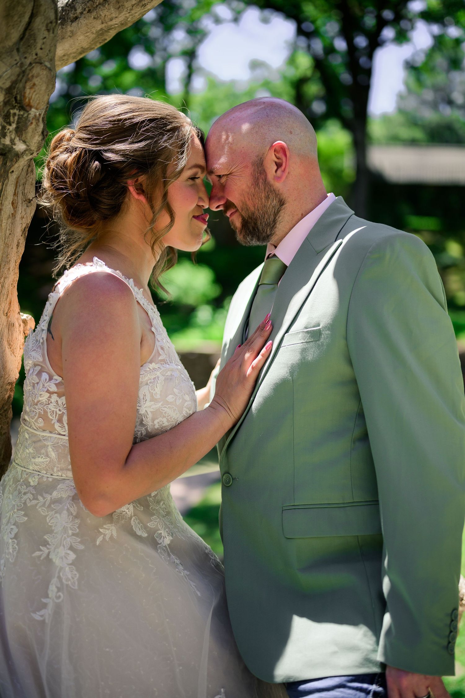 Bride and groom share an intimate forehead touch outdoors, she in lace gown, he in sage green suit.