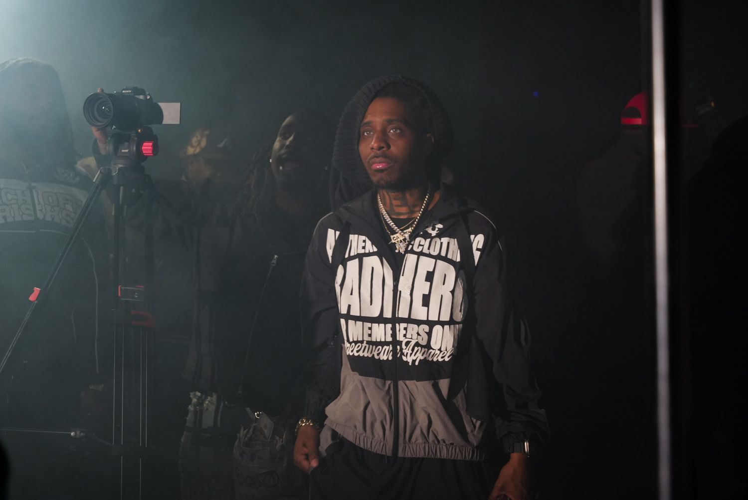 Male artist in graphic hoodie standing on dark stage under spotlight.