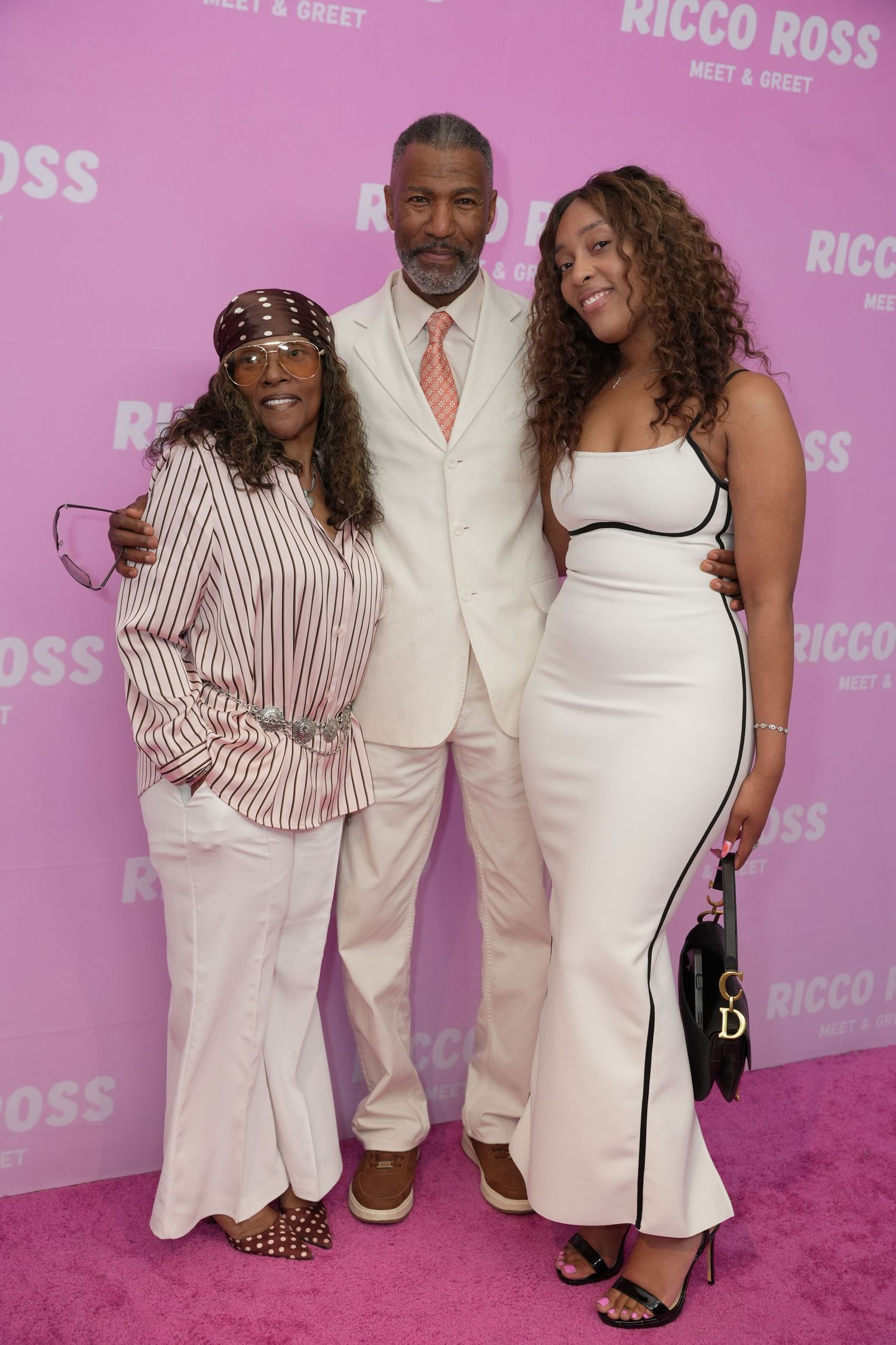 Three guests pose together on pink carpet at the Ricco Ross Meet and Greet, dressed in white and striped outfits.