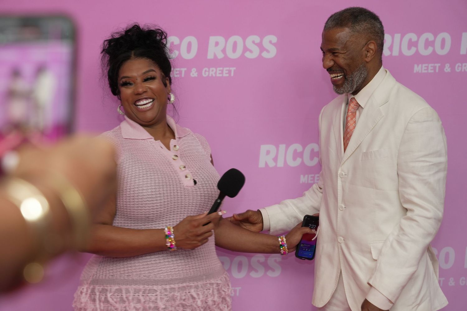 Woman in pink dress holds microphone while laughing with man in white suit at Ricco Ross Meet and Greet event.