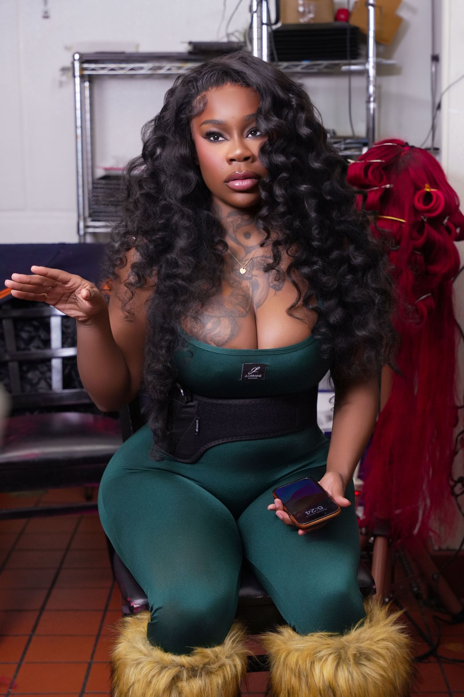 Woman in green outfit with waist trainer sitting and holding phone indoors.
