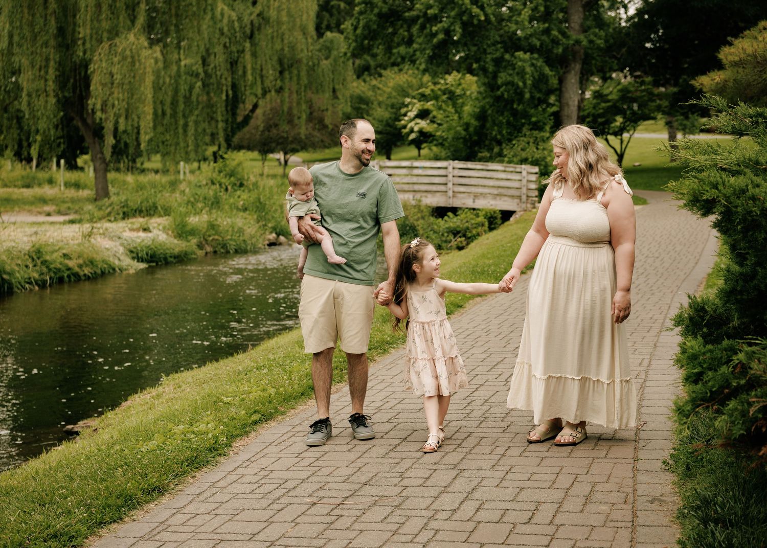 Family of four strolling along a scenic park pathway beside a pond, surrounded by lush green trees.