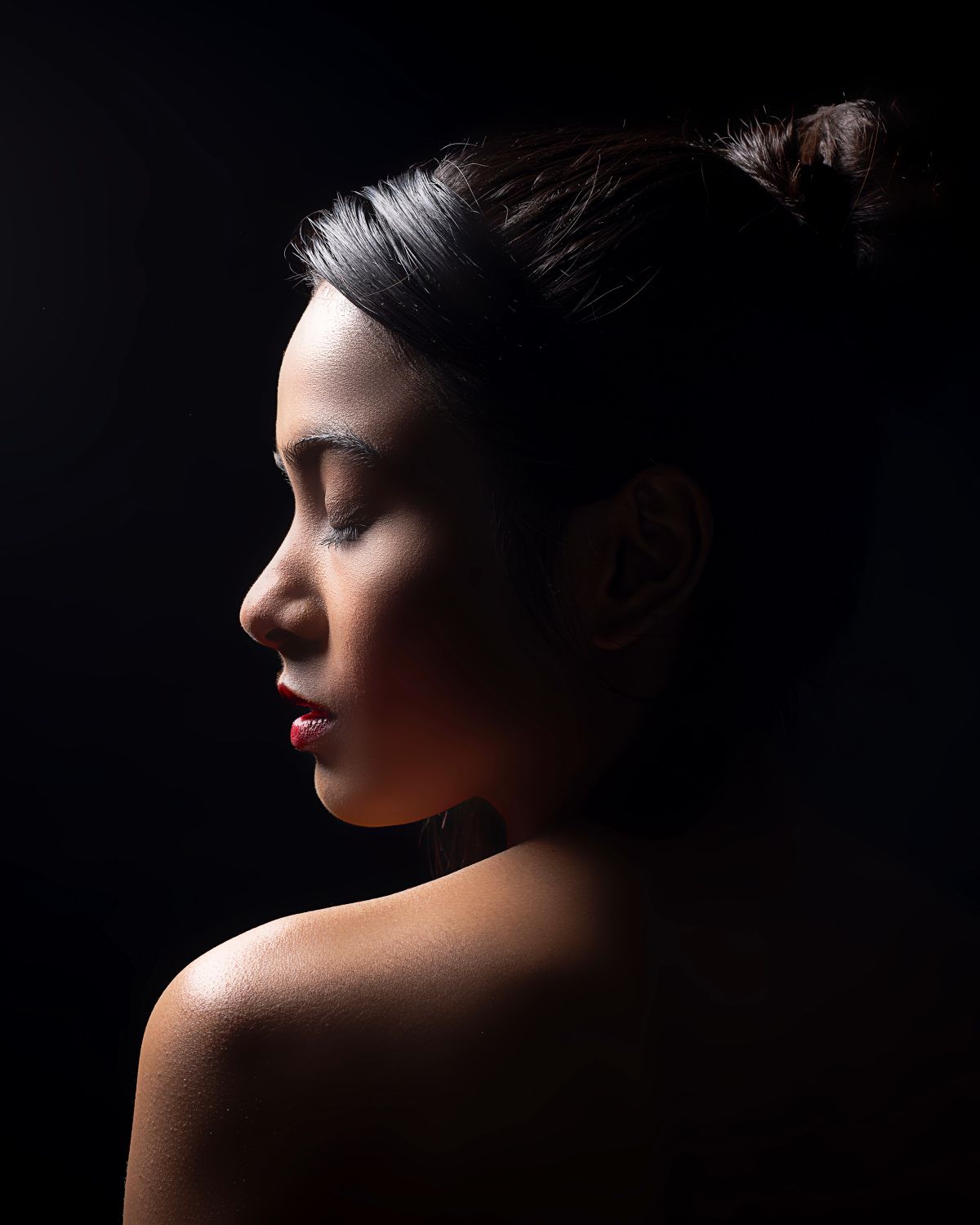 Profile portrait of a woman with dark hair in a bun, red lips, and bare shoulder against a black background.