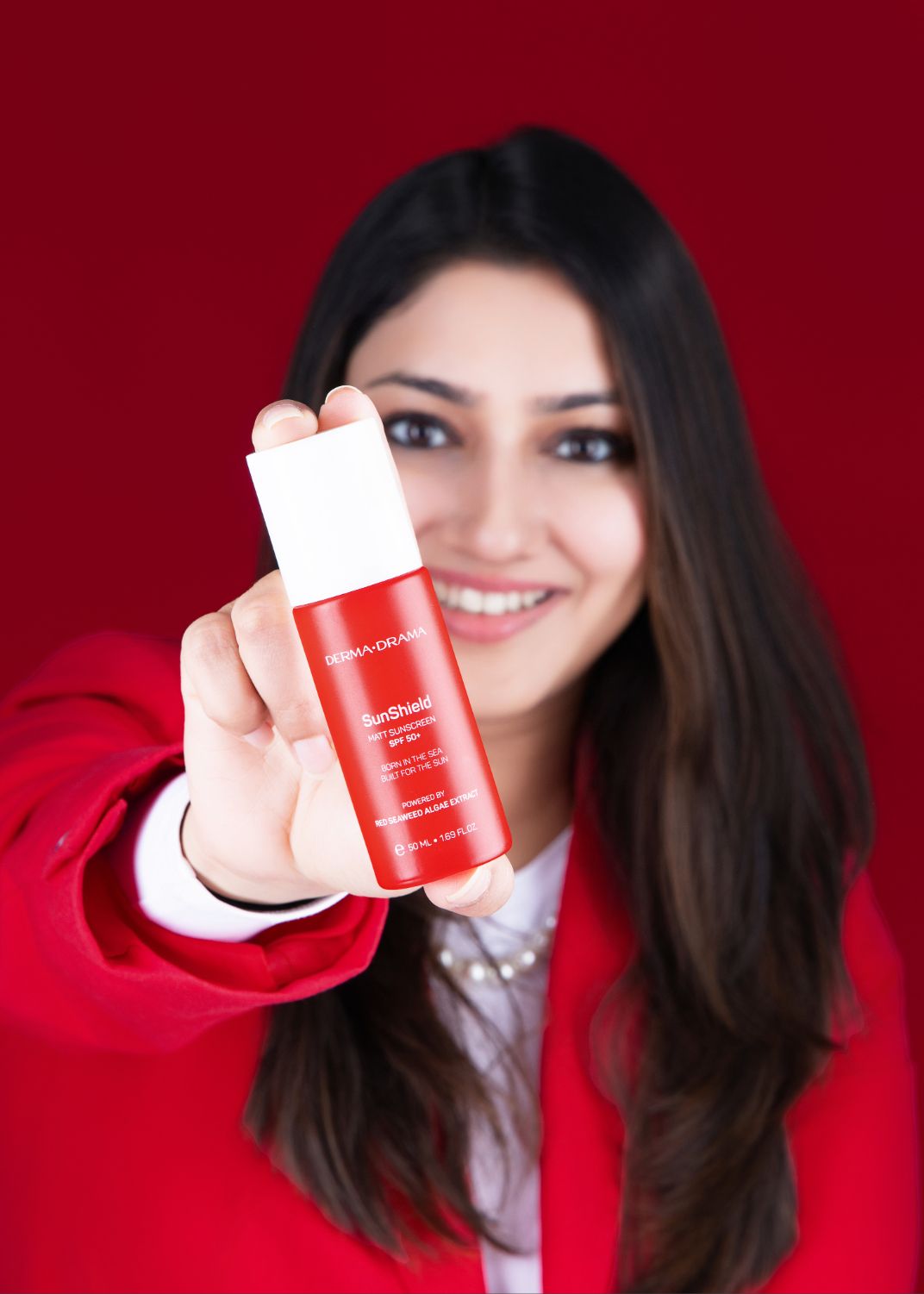 Person holding red skincare product bottle against burgundy background while wearing white and red clothing.