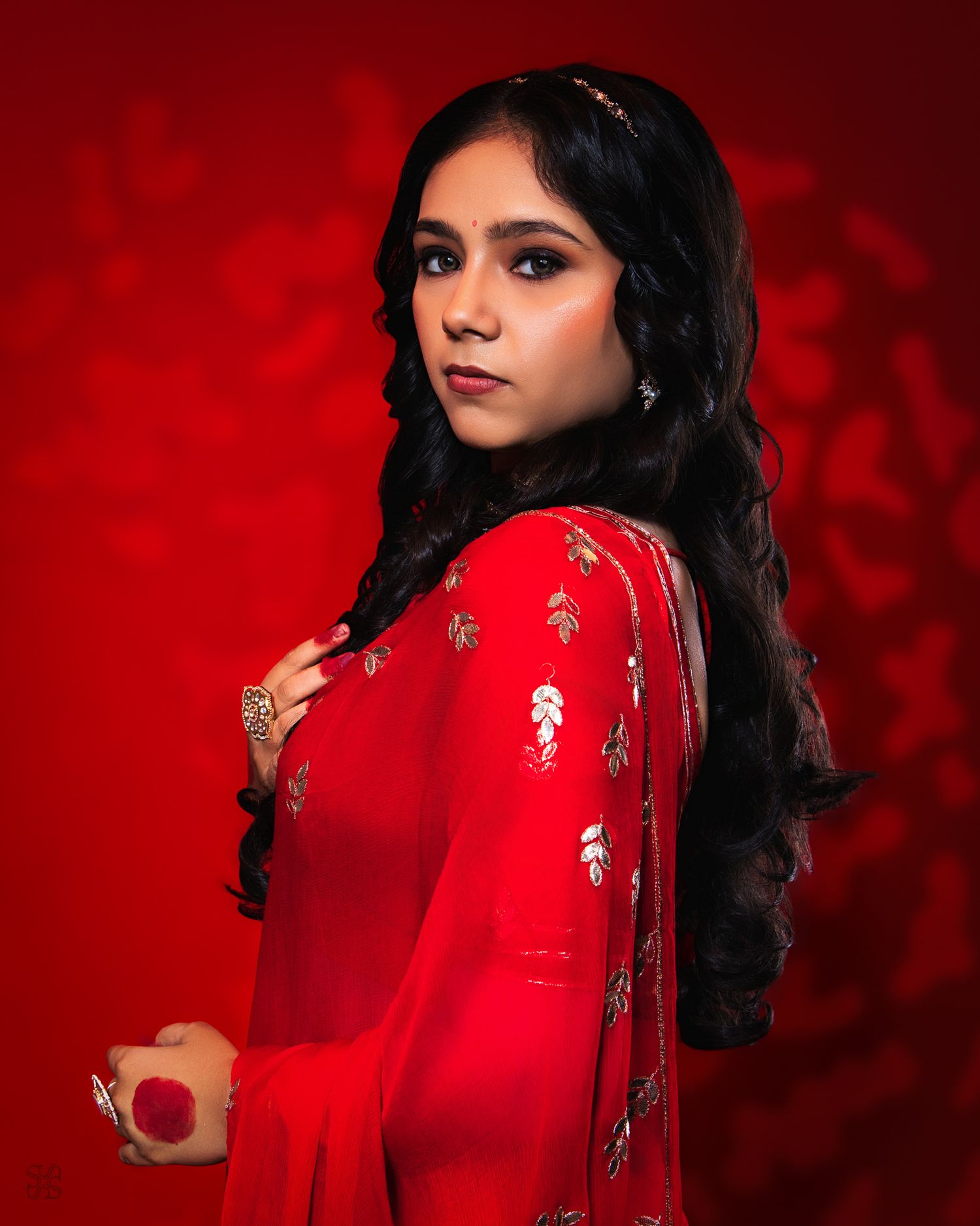 Young woman in red embroidered saree poses elegantly against a red background with dramatic lighting.