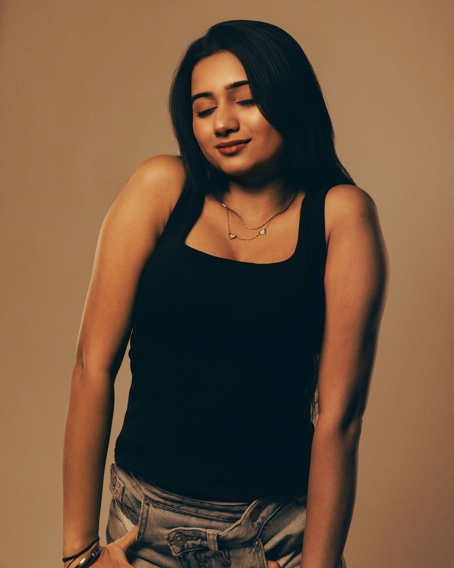 Young woman in black tank top and jeans posing confidently with a delicate necklace against a warm beige background.