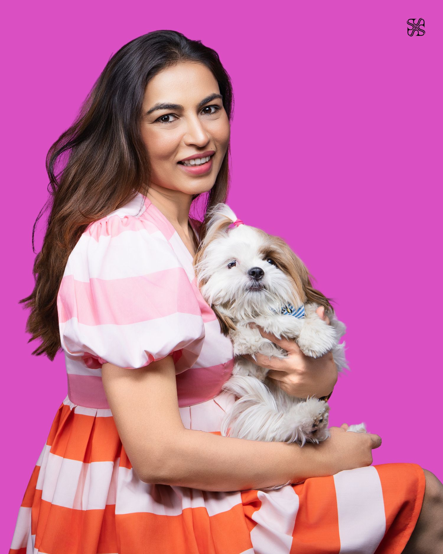 Woman in pink striped dress smiling and holding a small fluffy dog with a bow tie against a bright pink background.