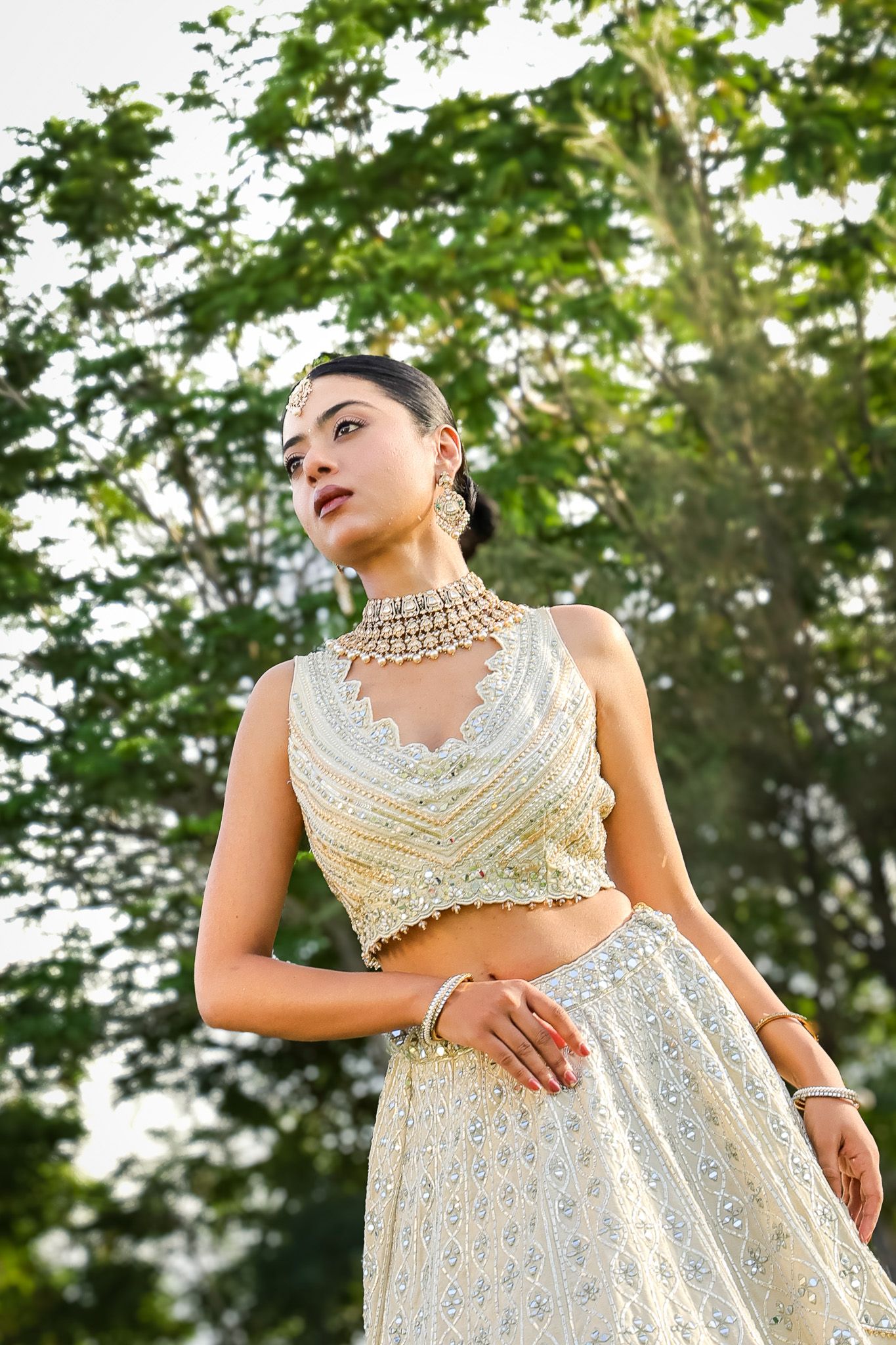Young woman in elegant white embroidered lehenga and kundan choker necklace poses outdoors with lush green trees.