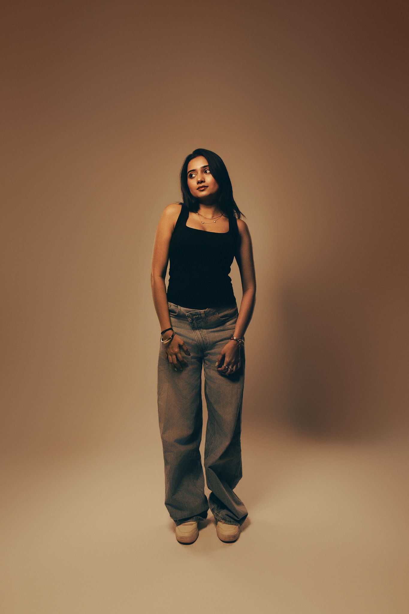 Young woman in black tank top and wide-leg gray pants posing against a warm beige studio background.